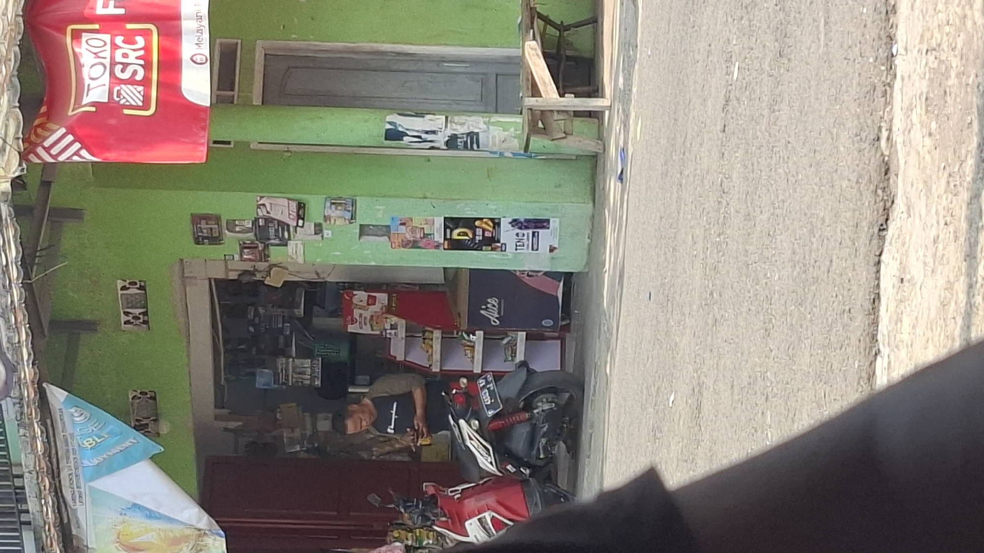 A street-level, medium-shot, vertical, full-color photograph shows the exterior of a small store in Cilaku, Indonesia. The store is painted lime green, with a red banner hanging above the entrance. The banner has white text that reads "TOKO SRC". To the left of the entrance is a red motorcycle, and to the right is a gray door. A man in a black t-shirt and gray sleeves is standing in the doorway, looking at his phone. Various posters and advertisements are affixed to the exterior walls of the store. A wooden bench is visible to the right of the door. The foreground shows a street with some debris on it.