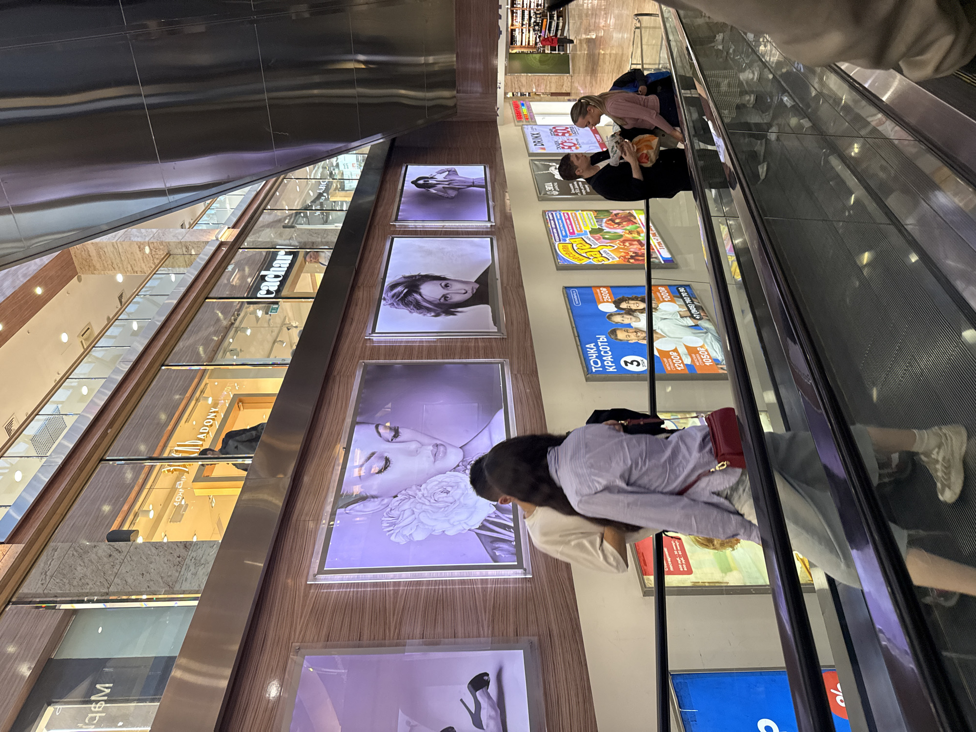 The image depicts the interior of a modern shopping mall, likely located in Moscow, Russia, judging by the Cyrillic text visible on advertisements. The scene is set on an escalator, with several individuals visible.
In the foreground, the metallic handrails and ribbed surface of the escalator are prominent. On the escalator, a woman with dark hair, wearing a light blue button-down shirt, a light-colored skirt, and white sneakers, is moving upwards. She has a red crossbody bag. To her left, a man with dark hair and a white t-shirt is partially visible, appearing to be in conversation with the woman.
Further up the escalator, another couple is interacting. A man in a black jacket and jeans is holding a plastic bag, and a woman with blonde hair in a pink sweater and dark pants is facing him.
Above the escalator, large illuminated advertisements are displayed on the wall. One features a close-up of a woman's face with flowers, and others show portraits of individuals. Storefronts are visible on the upper levels, with signage that includes Cyrillic lettering. The architecture of the mall is contemporary, with high ceilings and a reflective metal structure. The lighting suggests it is daytime.