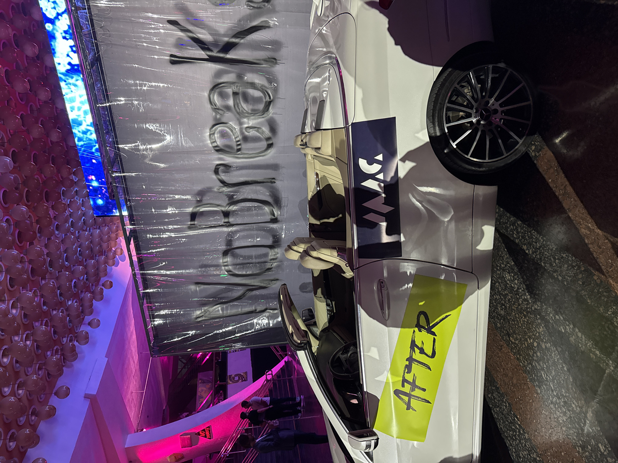 The image captures a car display in what appears to be an exhibition or event space, possibly in Yerevan, Armenia. A white car with an "AFTER" sticker on its side is a central element. The vehicle features a notable wheel design and is situated near signage that says "Before." In the background, there are a number of people, likely event attendees, and a wall with a decorative array of spherical objects, possibly balloons or light fixtures, adding to the ambiance. Adjacent to the wall is a display screen showing a blue and white pattern. The scene is well-lit, creating a vivid and engaging atmosphere.