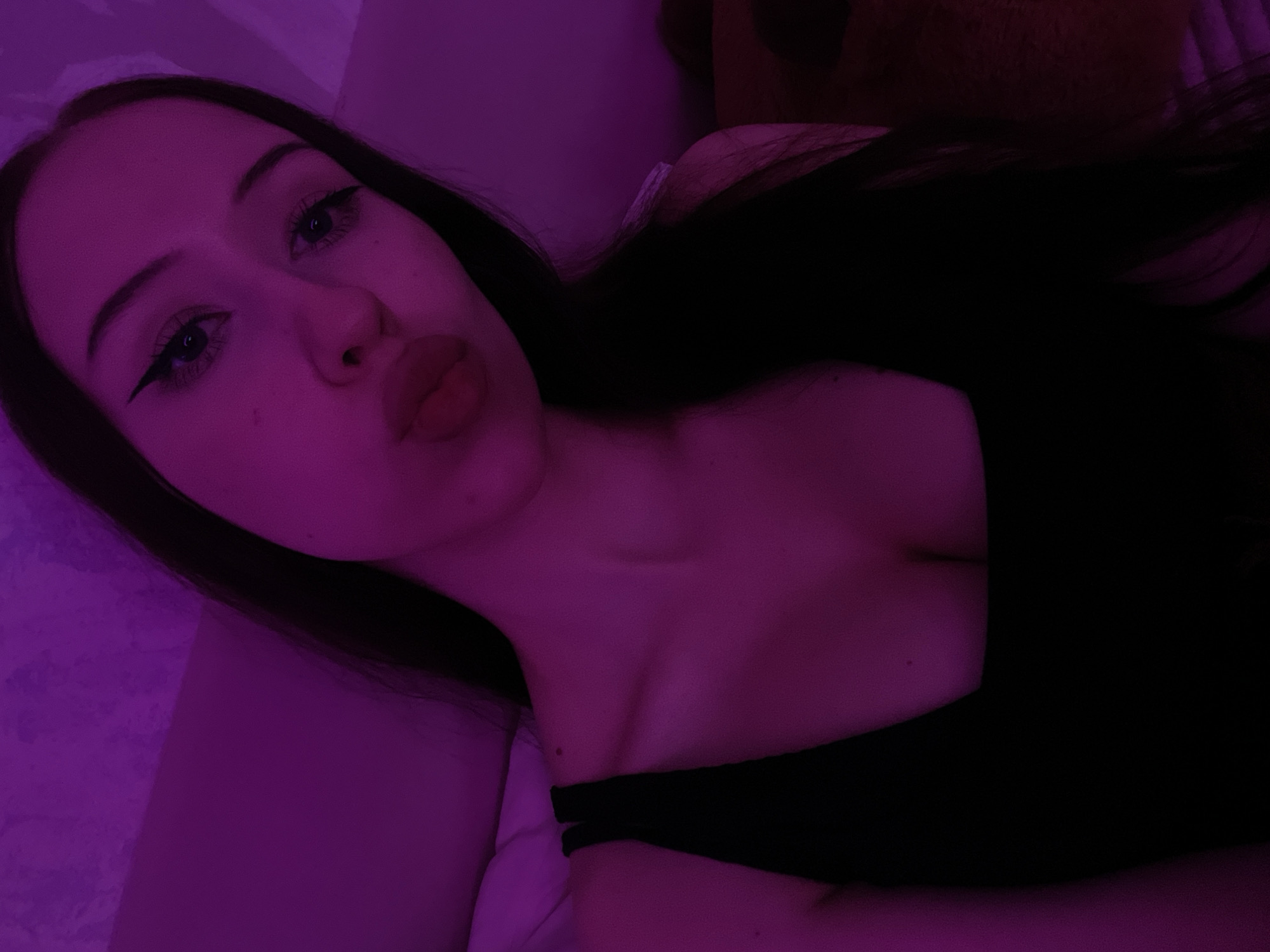 The image shows a close-up, selfie-style portrait of a young woman. She is looking directly at the camera with her lips pursed. The lighting is predominantly pink or purple, suggesting an artificial light source. She is wearing a black top with a square neckline, and her dark hair is visible around her face and shoulders. The background appears to be a wall with some texture and a hint of bedding or fabric. There are no discernible elements in the image that place the scene specifically in Votkinsk, Russia.