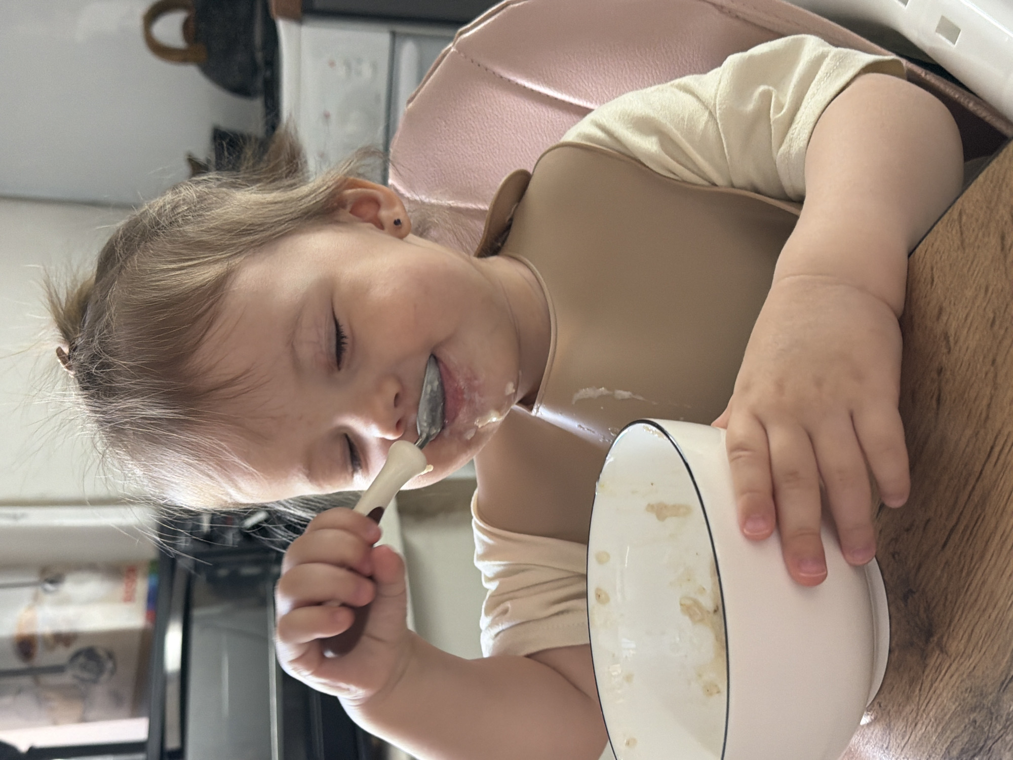 A young child with light brown hair is seated in a pink high chair, wearing a beige bib and a light-colored shirt. The child is holding a spoon in their right hand and is in the process of eating from a white bowl with a black rim. The bowl is partially empty, with remnants of food clinging to the sides. Food is also visible around the child's mouth. The child's eyes are closed or partially closed, suggesting concentration on the act of eating. The scene appears to be indoors, possibly in a kitchen or dining area, with a refrigerator or other appliance visible in the background. The location is not evident from the image, so the location in Reshetnikovo, Russia cannot be verified.