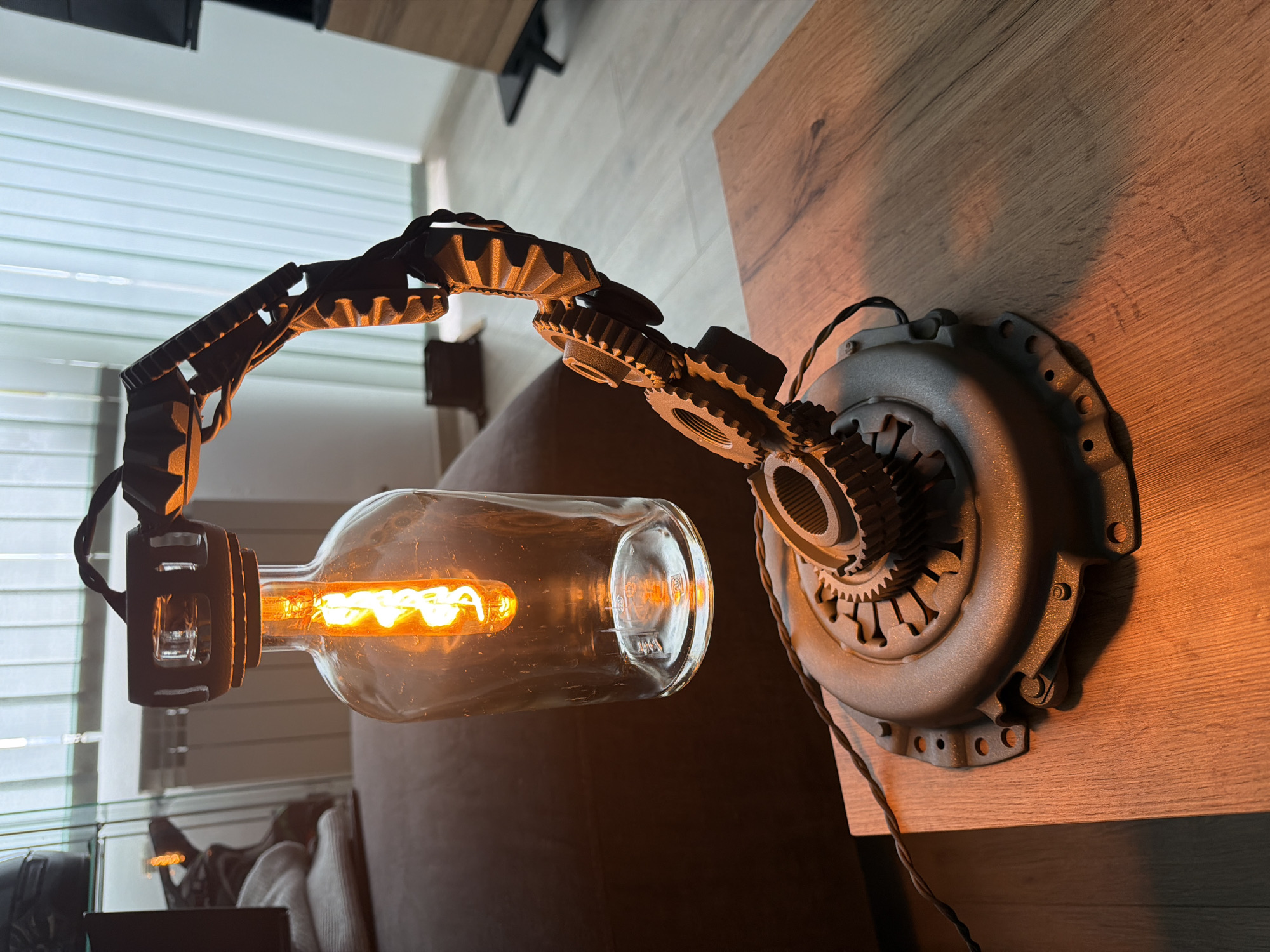 An industrial-style desk lamp or wall sconce, crafted from repurposed mechanical parts, is prominently featured in this image. The lamp's base is an automotive clutch pressure plate, mounted on a light brown wooden surface that displays a visible grain pattern. An articulated arm, composed of several interconnected gear-like segments with distinct teeth, extends from the base. The light source is an Edison-style filament bulb, which is housed inside a clear, elongated glass bottle, suggesting a repurposed item. The bulb emits a warm, orange glow, illuminating the immediate area.
The bottom of the glass bottle shows faint, molded text and symbols, including what appears to be a recycling emblem and partial letters like "JA". A dark electrical cord descends from the lamp's base along the side of the wooden surface. In the background, a window with white horizontal blinds or shutters is visible, providing a subtle light source. A dark, upholstered sofa or armchair is softly blurred in the background.
The setting is an indoor space, likely a living room or study, with the warm light from the lamp creating a cozy ambiance, indicative of evening or a low-light environment. No individuals are present, and no specific activity is taking place. The location is Sofia, Bulgaria.