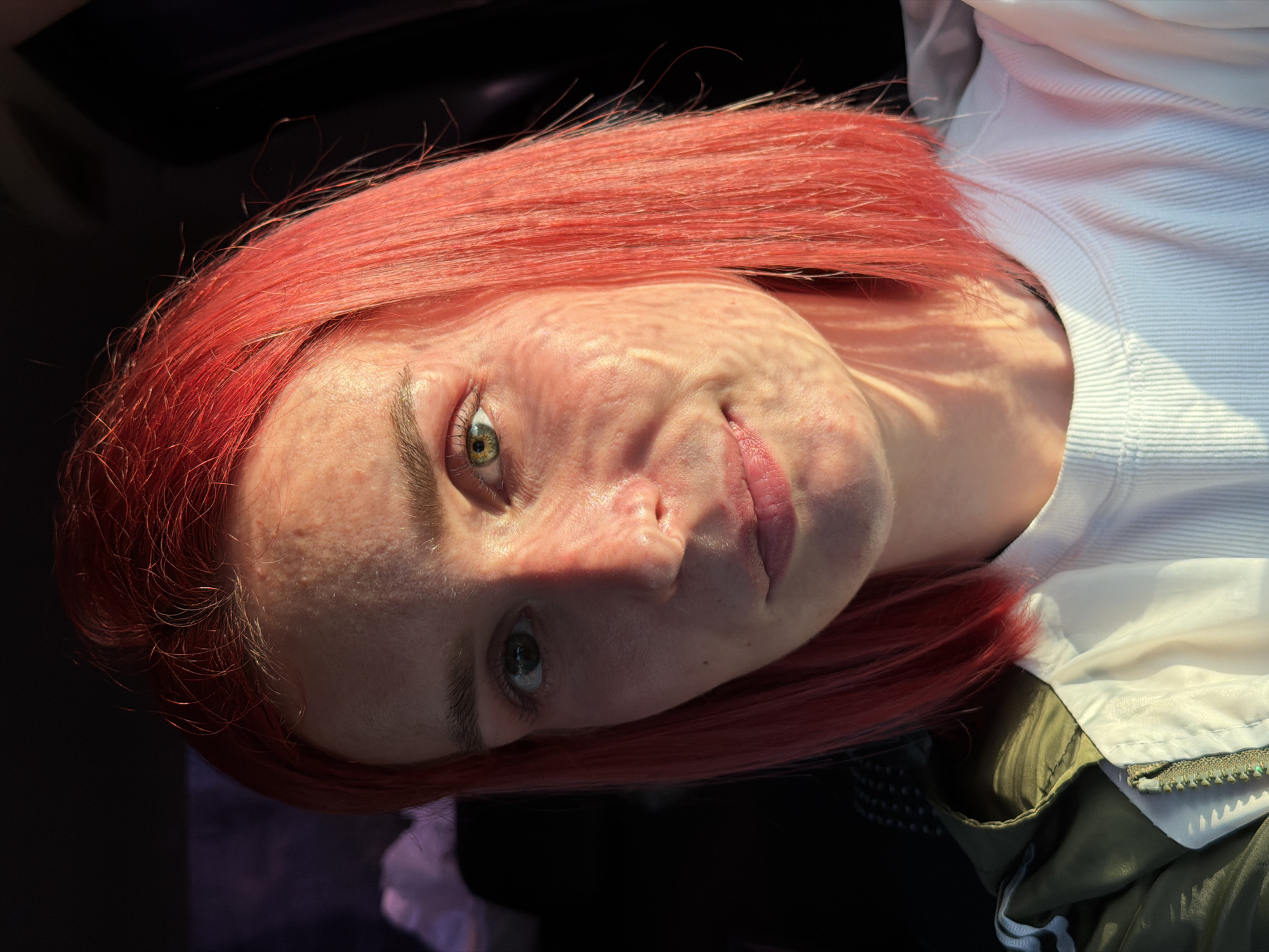In an outdoor, natural light setting, a young woman with bright red, shoulder-length hair is captured in a close-up, front-facing portrait. Her facial skin exhibits some texture and redness, with a distinct shadow cast across the left side of her face from a light source to her right. She has green eyes, and her lips are slightly parted in a subtle smile. She is wearing a white ribbed t-shirt and a green jacket with a zipper. The background is dark and out of focus, with hints of purple and white shapes suggesting a distant landscape or sky, possibly indicative of being in or near Chaikovsky, Russia, given the context provided, although this cannot be definitively confirmed from the image alone.