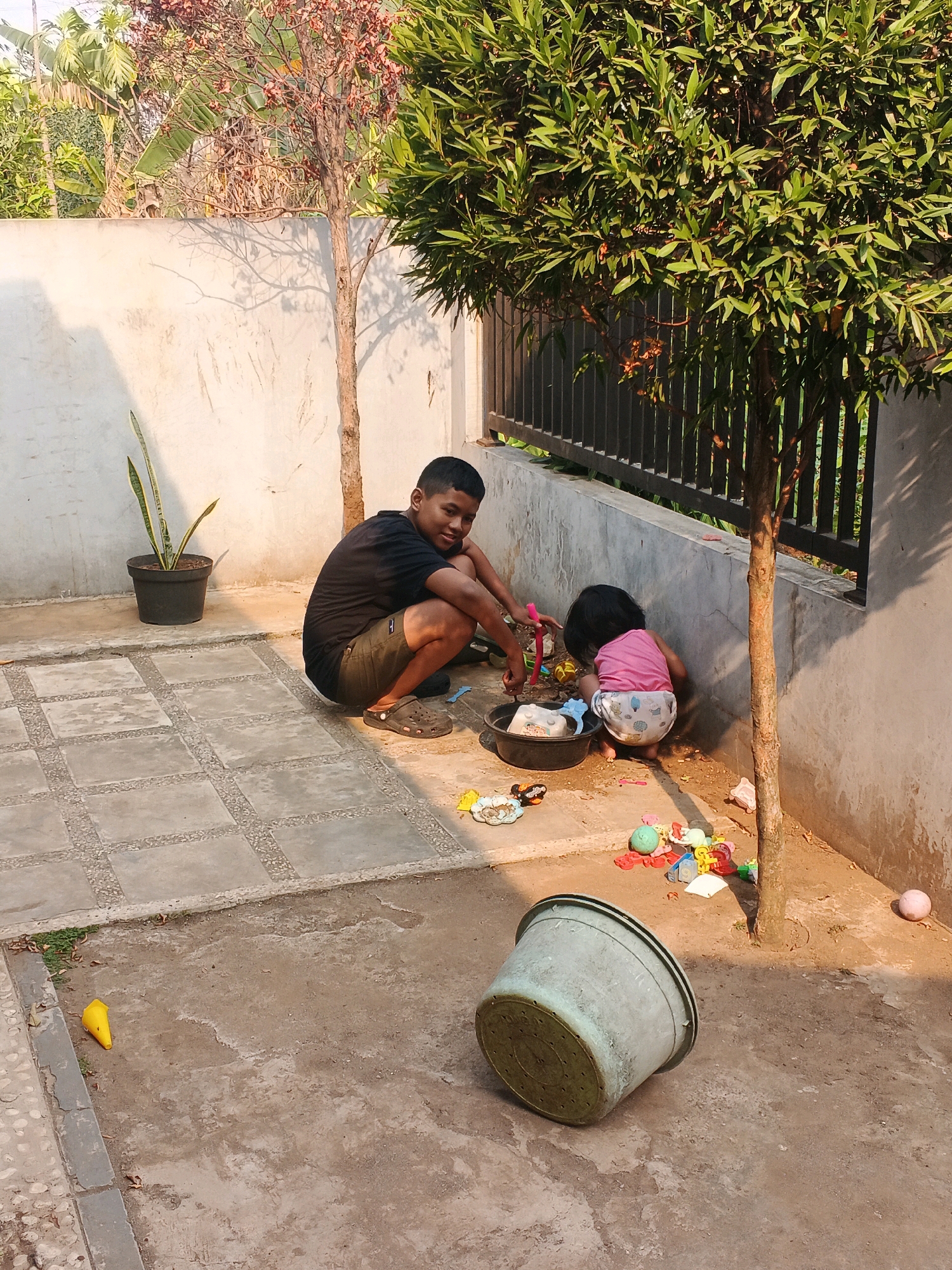 Two children are playing with toys in an outdoor area. A boy, appearing to be around 10-12 years old, is squatting on a paved surface, wearing a black t-shirt and shorts, and looking towards the camera with a smile. Next to him, a younger child, likely a toddler, is also squatting and playing with toys on the dirt ground, wearing a pink top and a diaper. Scattered around them are various colorful plastic toys, a black bucket, and a large light green bucket lying on its side in the foreground. The environment appears to be a backyard or patio, with a concrete wall behind the children and a small tree with dry leaves. The ground is a mix of concrete tiles and dirt. In the background, there are more trees and foliage, suggesting a location with greenery. The lighting indicates it is daytime, likely with direct sunlight casting shadows.