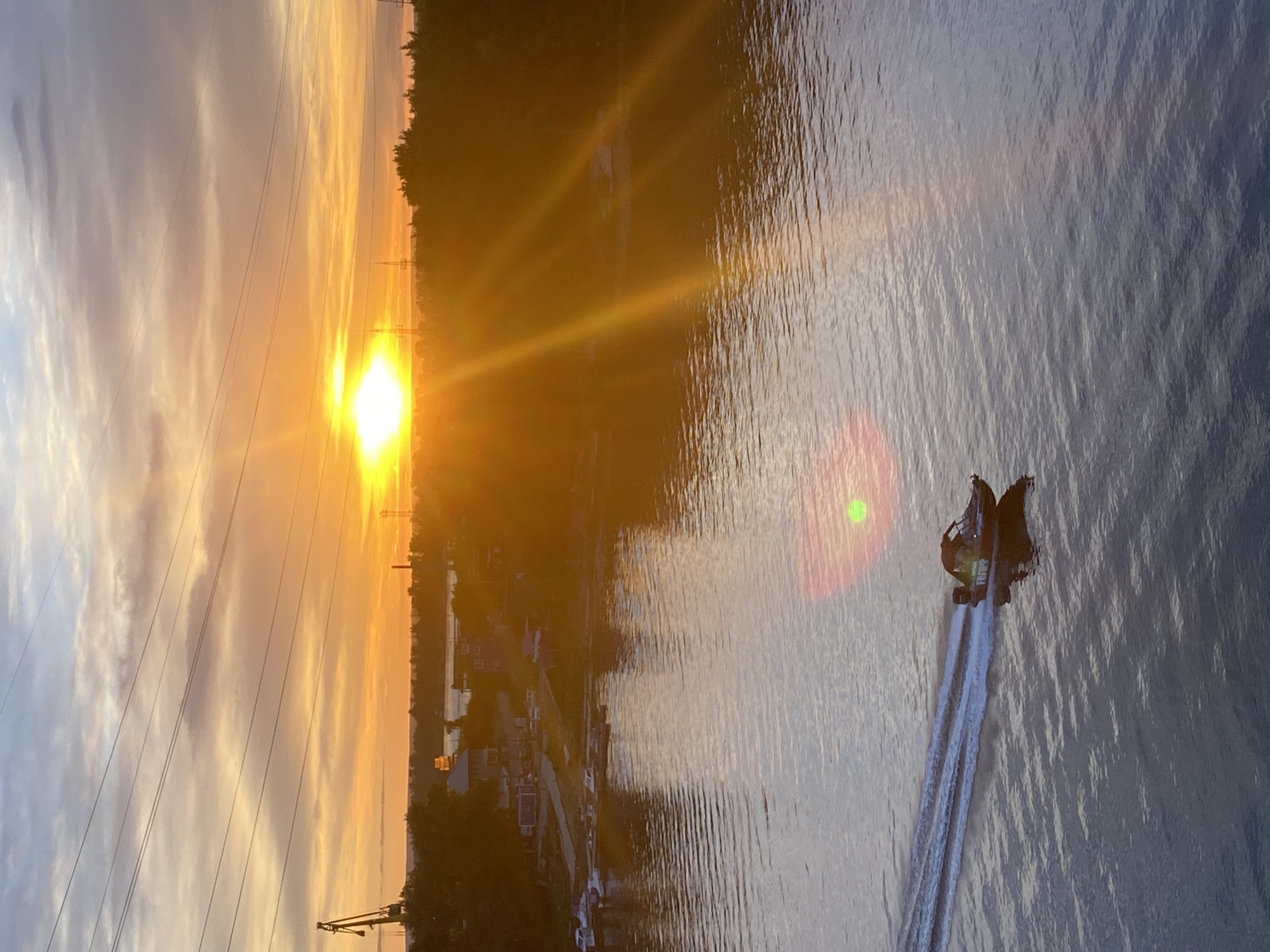 The image captures a serene waterscape at sunset, likely in Novokuybyshevsk, Russia. The sun, a brilliant orb of golden light, descends towards the horizon, casting a warm, orange glow across the sky and reflecting on the rippling water. Wispy clouds, tinged with the hues of dusk, add to the atmospheric beauty. Silhouetted against the vibrant sky are power lines and transmission towers, suggesting an industrial or semi-urban fringe.
In the foreground, a personal watercraft, possibly a jet ski, glides across the water, leaving a white trail in its wake. Two individuals are seated on the craft, their forms indistinct against the glare of the setting sun. The water's surface is textured with light and shadow, creating an impression of gentle movement. The overall mood is peaceful and contemplative, evoking the end of a day and the quiet beauty of nature illuminated by the fading sunlight. A faint green lens flare is visible in the water, indicating the bright light source.