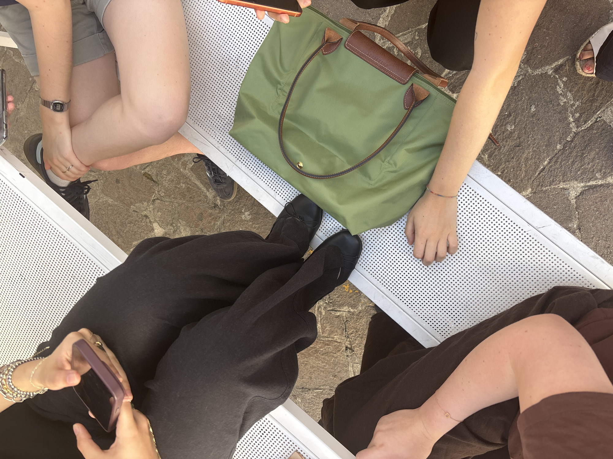 The overhead shot shows a group of people seated on a perforated white bench outdoors, with a cobblestone ground visible below. The people are partially cropped, with only their legs, arms, and torsos visible. One person on the left is wearing black pants and holding a purple-cased smartphone. Next to them, another person is wearing shorts and a watch, with their legs crossed. In front of them is a large olive green tote bag with brown leather accents. A third person on the right is wearing brown clothing and has a thin bracelet on their wrist. A fourth person's arm is visible on the far right, holding an orange-cased smartphone. The bench appears to be in an urban setting, possibly a public square or park area in Milan, Italy, given the cobblestone paving.
