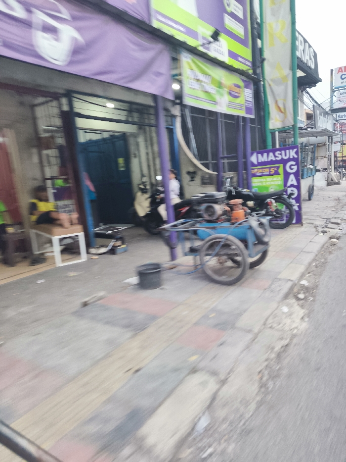 The scene is a street in Cileunyi, Indonesia, located at coordinates -6.933 latitude, 107.724 longitude. A blue handcart with a large, black wheel is parked on the sidewalk. A man sits on a bench, and a second man is visible, walking towards the cart. A motorcycle with a sidecar is also present in the background. The scene takes place in front of a building with a purple awning and a sign that reads "MASUK GAP". The street is paved with grey concrete and has a slight incline. The sky is overcast. The sidewalk is cluttered with debris.