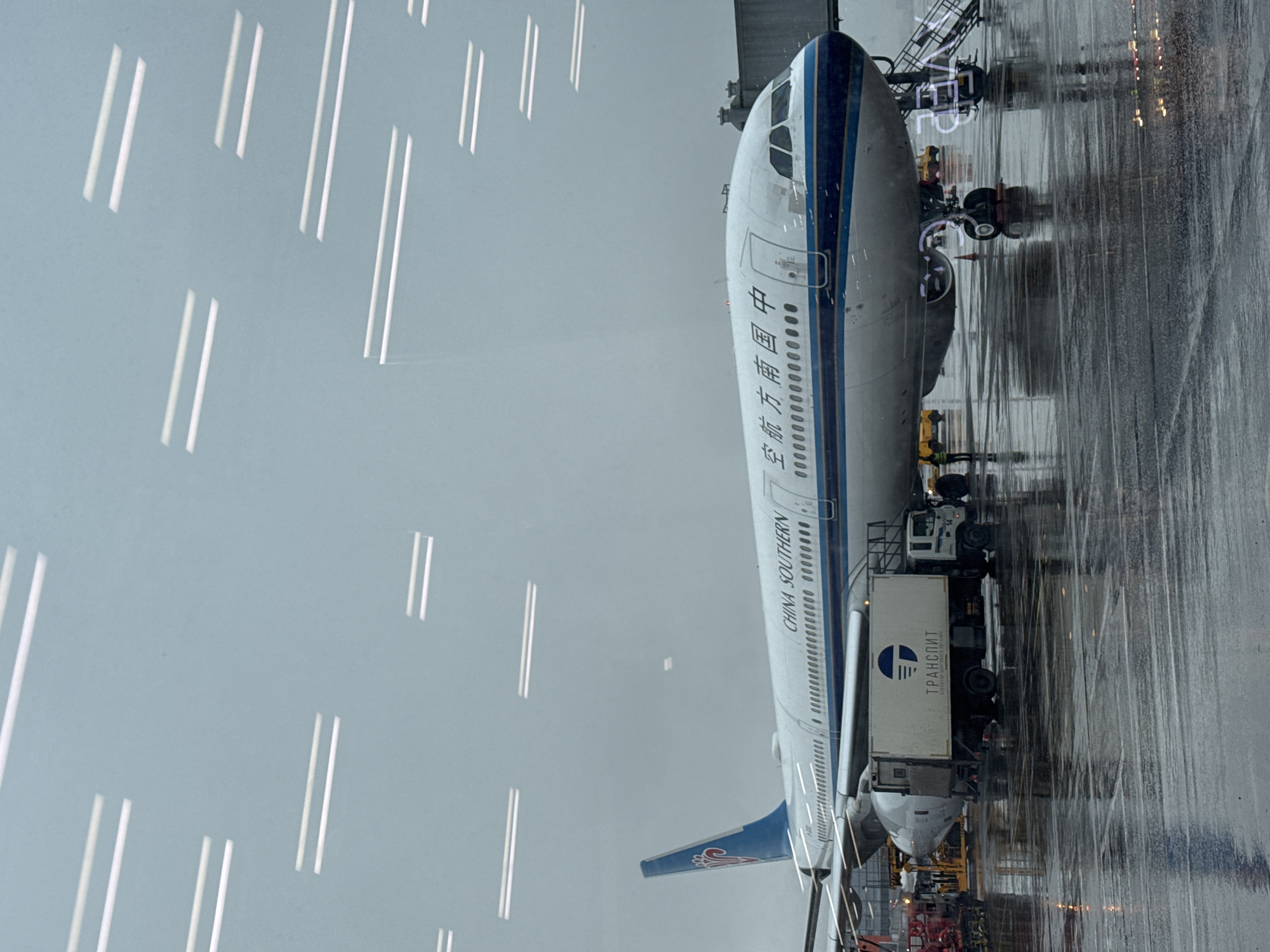 A China Southern airplane is parked at an airport in Khimki, Russia. The location is identifiable from the Cyrillic text "ТРАНСПИТ" visible on the side of a cargo container servicing the aircraft. The scene appears to be in rainy conditions. Parallel white lines are on the left side of the image, and airport infrastructure is visible near the plane.