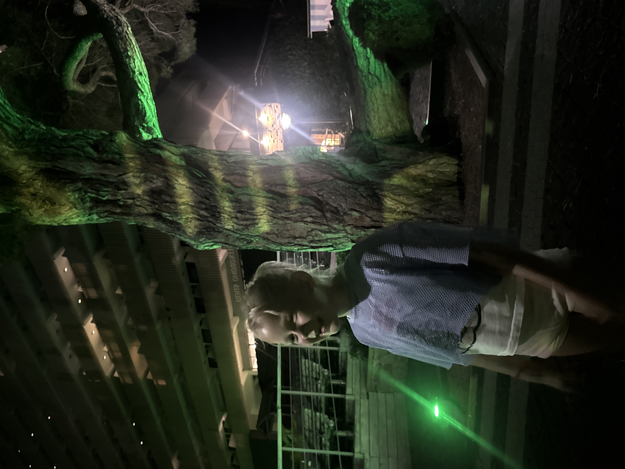 The image depicts a young blonde girl standing near a large tree illuminated by green lights. The girl is wearing a blue mesh top over a white undershirt and light-colored shorts. She is smiling and looking to her left. Behind her, a multi-story building with balconies and lights is visible. The setting appears to be outdoors at night, possibly in a park or resort area. The text "ЦЕНТР ОМОЛОЖ" is visible on the building, which translates to "Center for Rejuvenation" in Russian. Based on the visible text and the geographical context provided, this scene is located in Yalta, Ukraine.