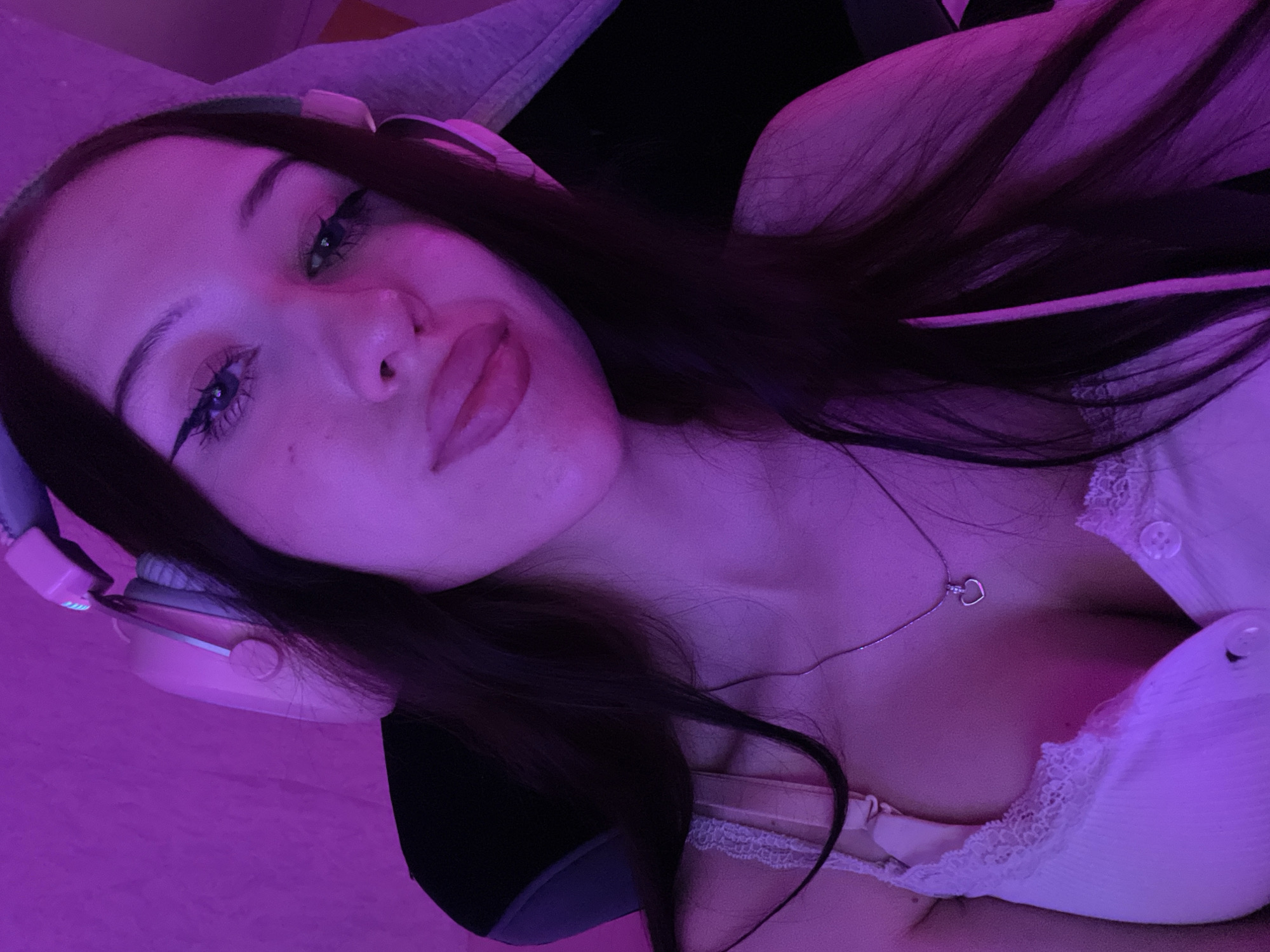 A young woman with long, dark hair is shown from the chest up, wearing a light-colored bra with lace trim and a silver necklace with a heart pendant. She has headphones with pink accents over her ears. The scene is illuminated by purple lighting, creating a vibrant and moody atmosphere. The background is a solid pink wall. The woman is looking towards the camera with a direct gaze, her eyes are accentuated with winged eyeliner. Her lips are full and slightly parted. The overall impression is a close-up, selfie-style portrait. There is no indication of location within Votkinsk, Russia.