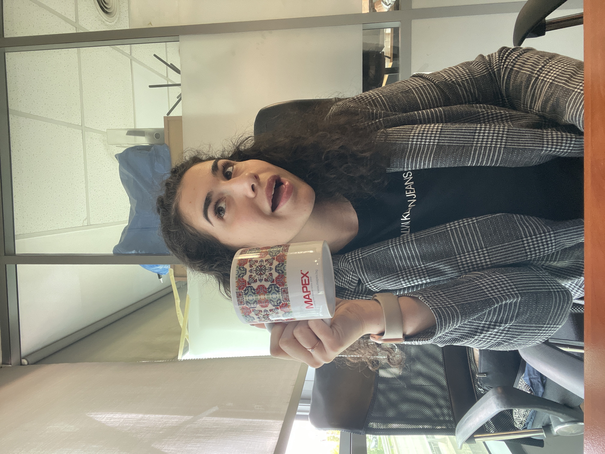 A young woman is holding a "MAPEX" mug. She is wearing a black Calvin Klein Jeans shirt and a black-and-white plaid blazer. The scene appears to be an office setting with a ceiling vent and blinds in the background. It could be Sofia, Bulgaria due to the fact the mug mentions "www.mapex.by" and it could be a Bulgarian company, but I am not completely sure.