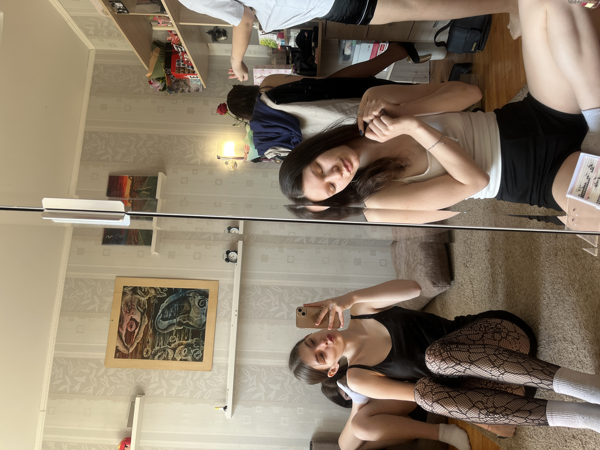 A mirror selfie in a room with floral patterned wallpaper, likely Saint Petersburg, Russia, based on architectural styles and interior design, displays two individuals. One person, wearing a black tank top and fishnet stockings, lies on a carpeted floor, holding a smartphone taking a selfie. Their reflection shows a white top and black shorts. A second individual is standing, partially visible at the top of the frame, wearing a white t-shirt and black shorts. Background details include a shelf stocked with various items, pictures on the wall, a bed, and furniture. A red rose is placed near the bed.