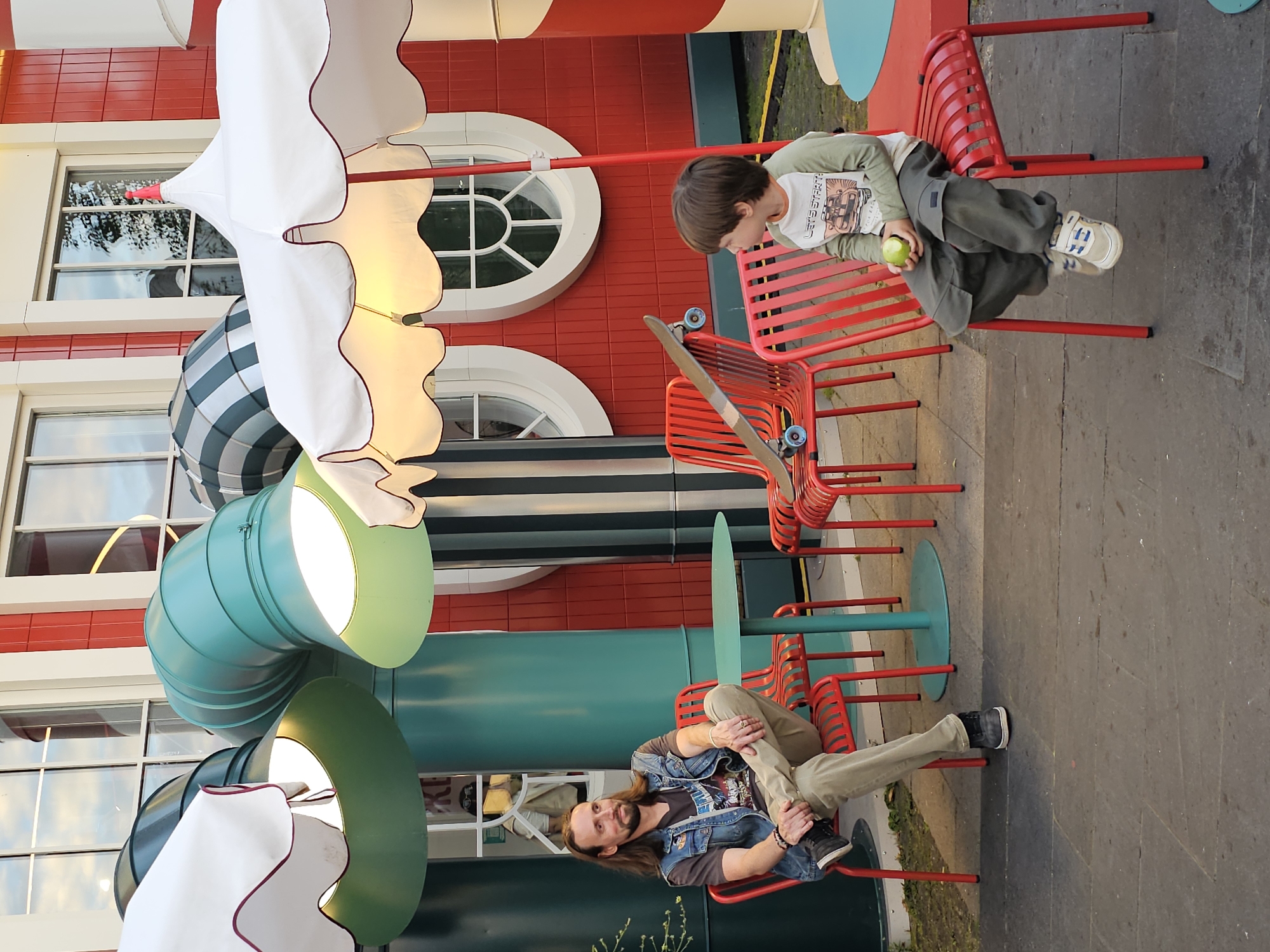 A young boy and a man are sitting on red chairs outside a building with red siding and a green, twisted tube structure. The boy is wearing a long-sleeved shirt and pants, and he is holding a green apple in his hand. A skateboard rests on the chair next to him. The man has long brown hair and a beard, and he is wearing a denim vest over a t-shirt and pants. He is looking towards the camera with a slight smile. The setting appears to be a public outdoor space, possibly a park or entertainment venue, given the colorful architecture and seating. The lighting suggests it is daytime. The mood appears relaxed and casual.