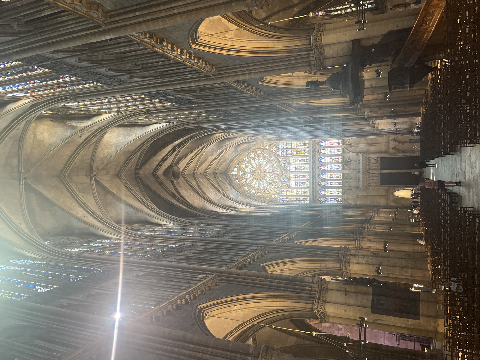 The image shows the interior of Metz Cathedral, located in Metz, France. The cathedral features Gothic architecture with ribbed vaults, stained glass windows, and rows of chairs in the nave. A large rose window is visible at the far end of the nave, with sunlight streaming through the stained glass. Several people are walking towards the back of the cathedral on the right side of the image.