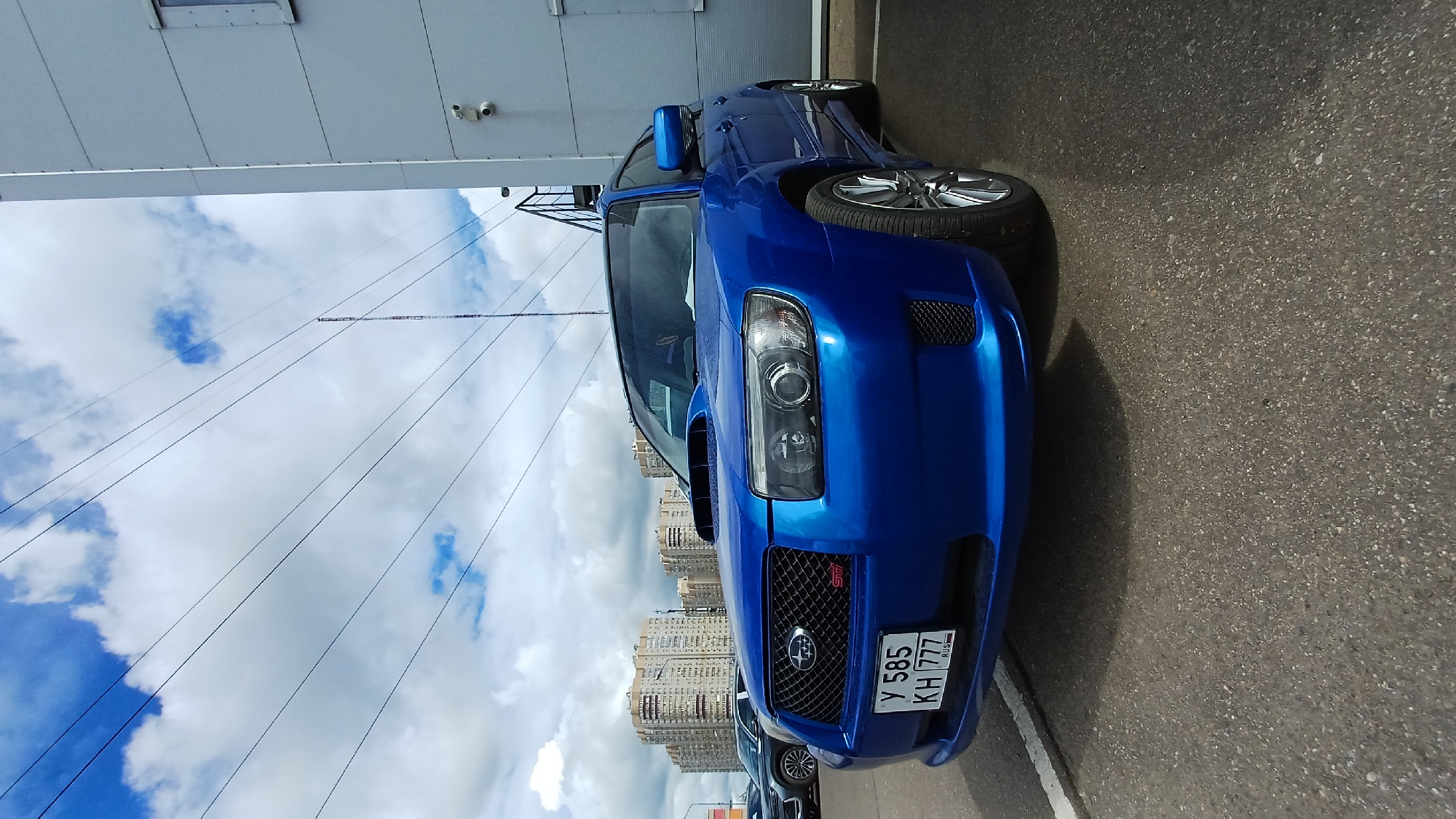 A blue Subaru Forester STI is parked on asphalt, facing the camera. The license plate reads "Y 585 KH 777 RUS". The car is parked in a city setting. In the background are multi-story residential buildings. Overhead are clouds and overhead electrical wires. The photo was taken near Balashikha, Russia. A building is visible to the left.