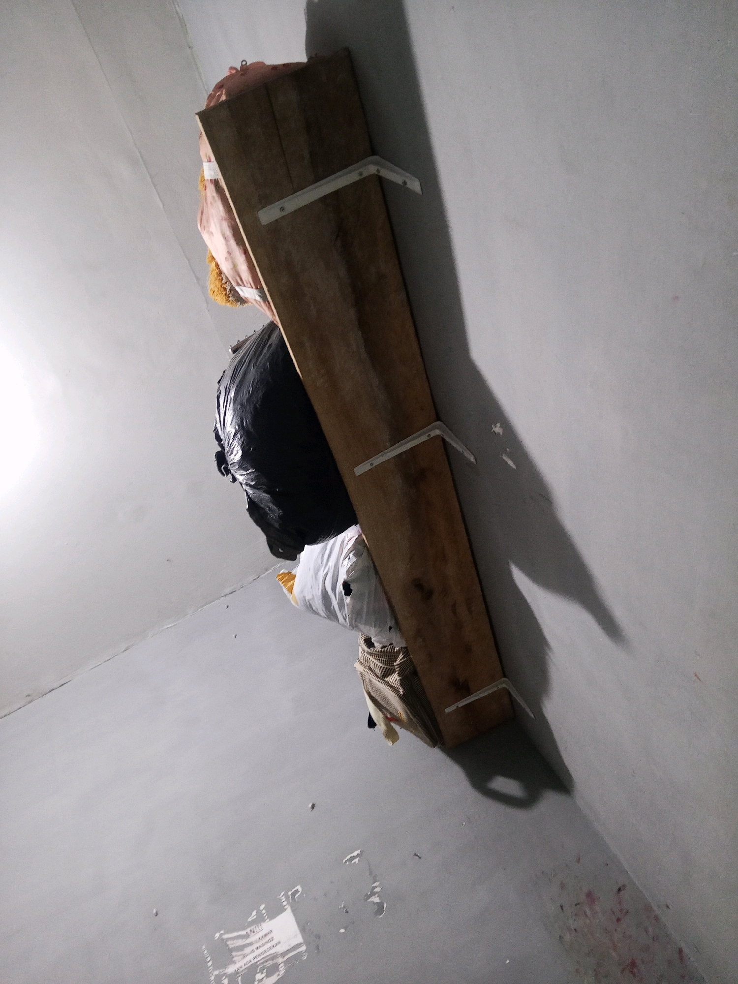 A raw wooden plank, supported by two white metal brackets, is affixed to a light gray concrete wall at an angle. The plank serves as a shelf, holding several items. At the top, a pinkish-orange bag with a yellow fringe is visible, followed by a large black plastic bag. Beneath that, a white bag with yellow trim is partially obscured, and at the bottom, a patterned fabric item is visible. The overall environment appears to be an interior space, possibly a room or storage area, with a plain concrete floor and walls. The lighting suggests an indoor setting with artificial light. No individuals are present in the image. The location of Lubuklinggau, Indonesia, is not discernable from the visual information provided in the image.