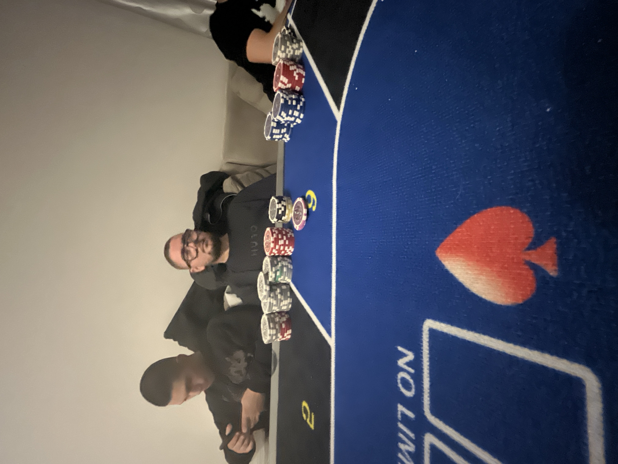 Two men are sitting at a blue poker table. There are stacks of poker chips in front of them. The poker table has a spade design on it and the words "No Limit". The photo appears to be taken inside, likely a home, with a plain white wall in the background. The location is not discernible to Burgas, Bulgaria, without additional information.