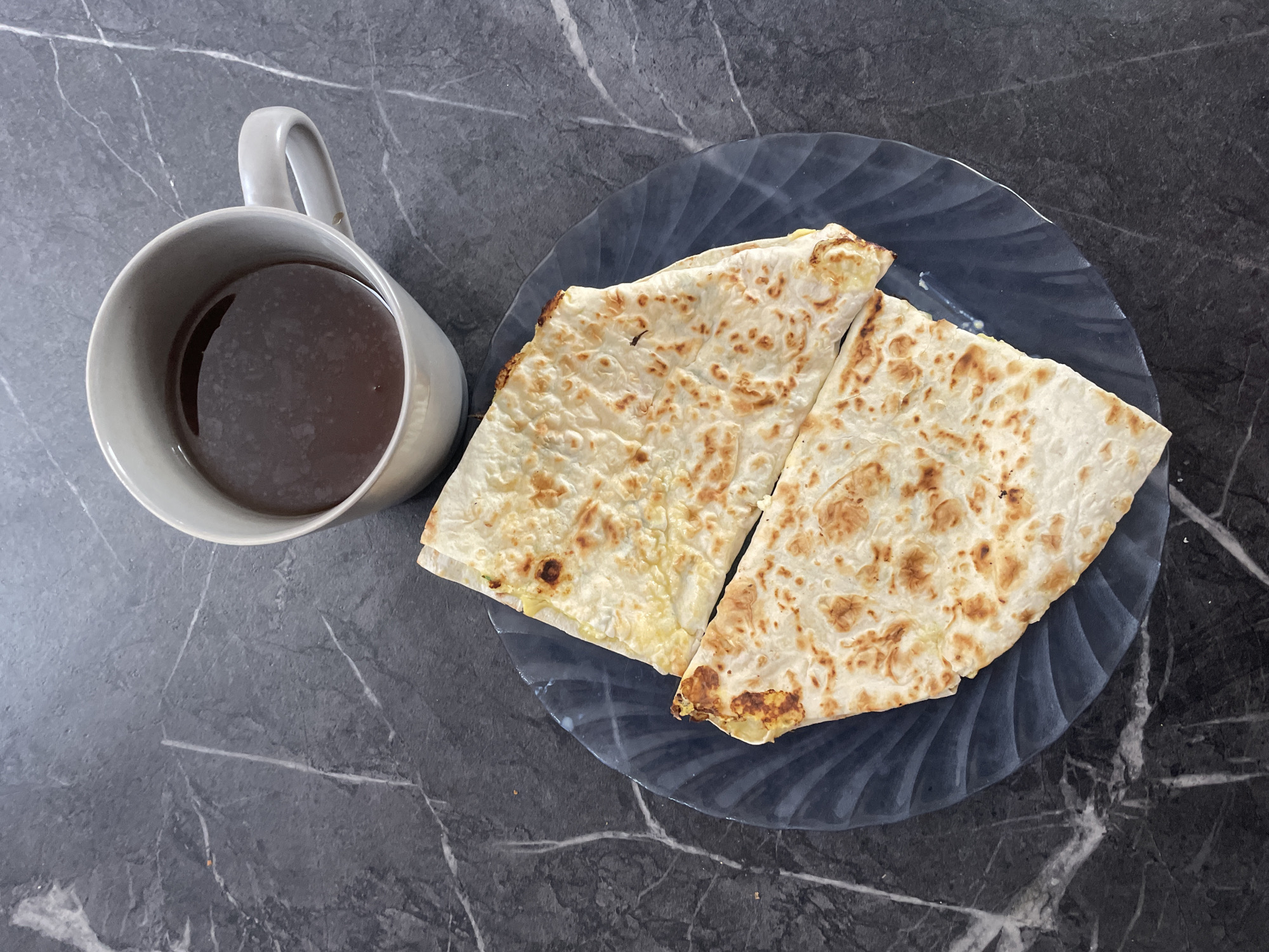 A top-down shot captures a cup of dark liquid, possibly coffee or tea, beside a plate of quesadillas on a dark, marbled surface. The cup is light gray with a prominent handle. The plate has a wavy pattern and is dark gray or blue. The quesadillas are cut into two pieces, showing golden-brown grill marks. The background is the dark, marbled surface with visible veins. The scene appears to be a breakfast or snack setting. There is no information to suggest this is in the city of Baranovka, Russia.