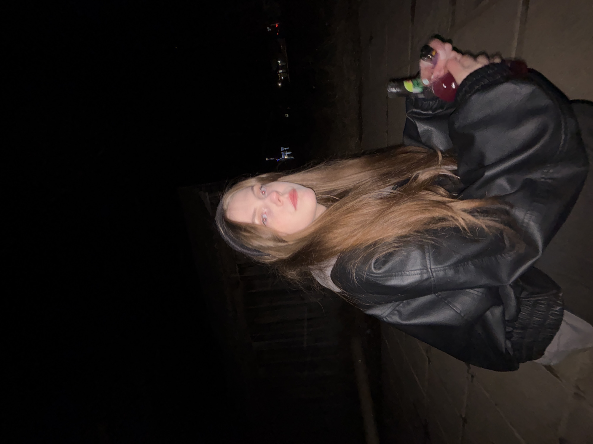 A young woman with long brown hair and fair skin is holding a bottle with a dark red liquid in it. She is wearing a black leather jacket. The image appears to be taken at night, with dark surroundings. In the background, there are some lights that suggest the location might be in the city of Фоки, Russia.