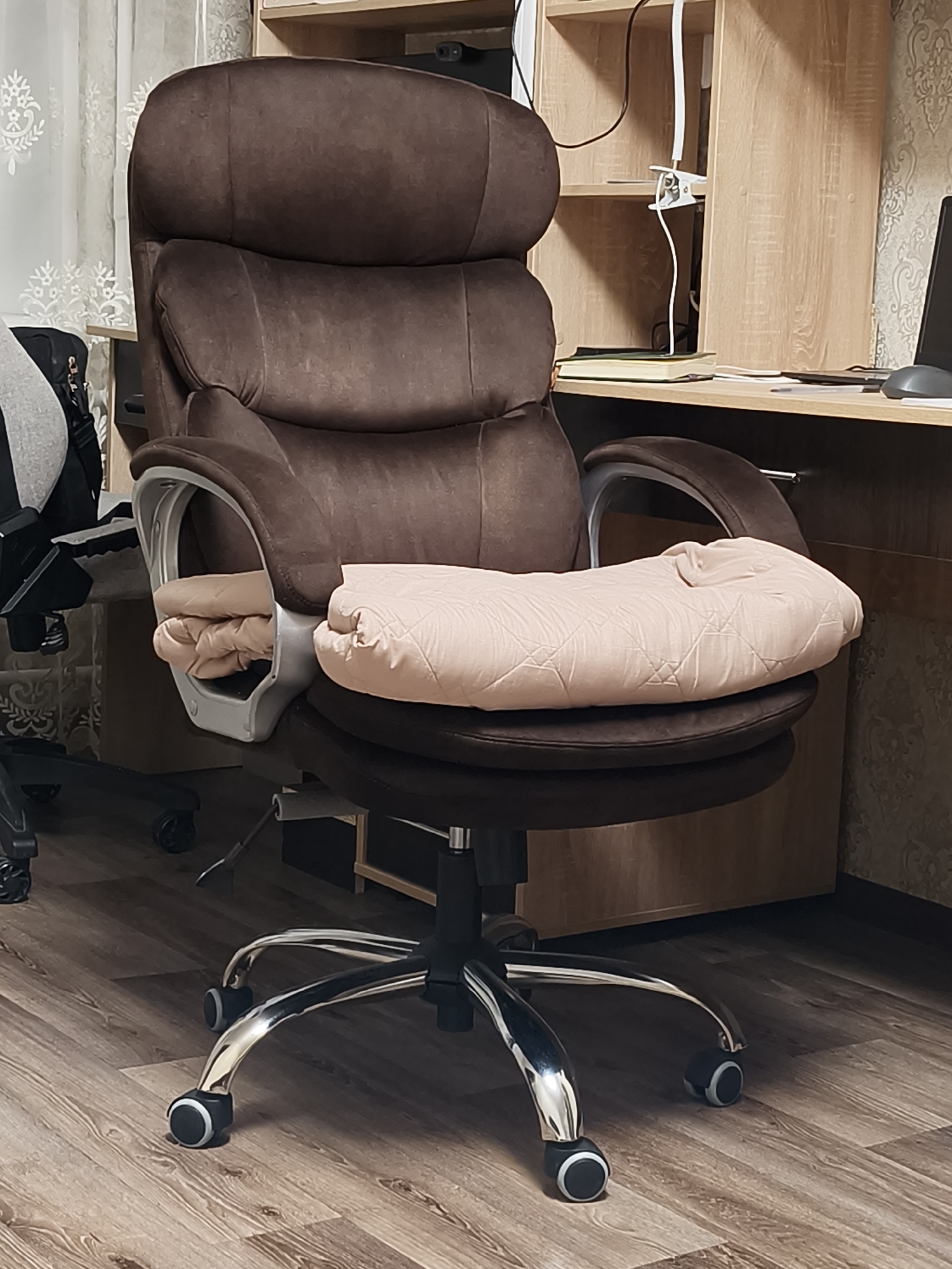 A close-up shot features a brown office chair with a high back, situated on a light brown wood-patterned floor. A folded, quilted light peach-colored blanket is draped on the chair's seat. The chair has a chrome base with black casters and chrome armrests. To the left, part of another black office chair is visible. Behind the brown chair, there is a wooden desk and a wooden cabinet with a light brown wood grain pattern. A computer monitor can be seen on the desk. The background includes a curtain on the left and patterned wallpaper on the right. The location cannot be determined based on the image.