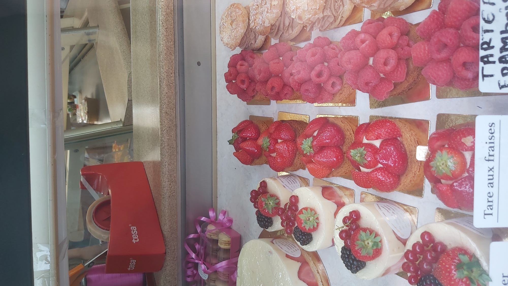 The image displays a selection of French pastries in a refrigerated display case, likely in a boulangerie or pâtisserie. In the foreground, various tarts and cakes adorned with fresh strawberries, raspberries, red currants, and blackberries are arranged. One tart in the lower foreground has a label that reads "Tarte aux fraises" (Strawberry tart). To the left, a large round cake with white frosting and strawberries is visible. In the background, behind the pastries, a red Tesa tape dispenser sits on the counter. The display case appears to be made of glass, allowing a clear view of the products. The overall environment suggests a commercial setting focused on baked goods. There are no people or animals present in the image. The lighting suggests an indoor commercial environment. The presence of "Tarte aux fraises" suggests the location is in France, likely Paris given the commonality of such establishments there.