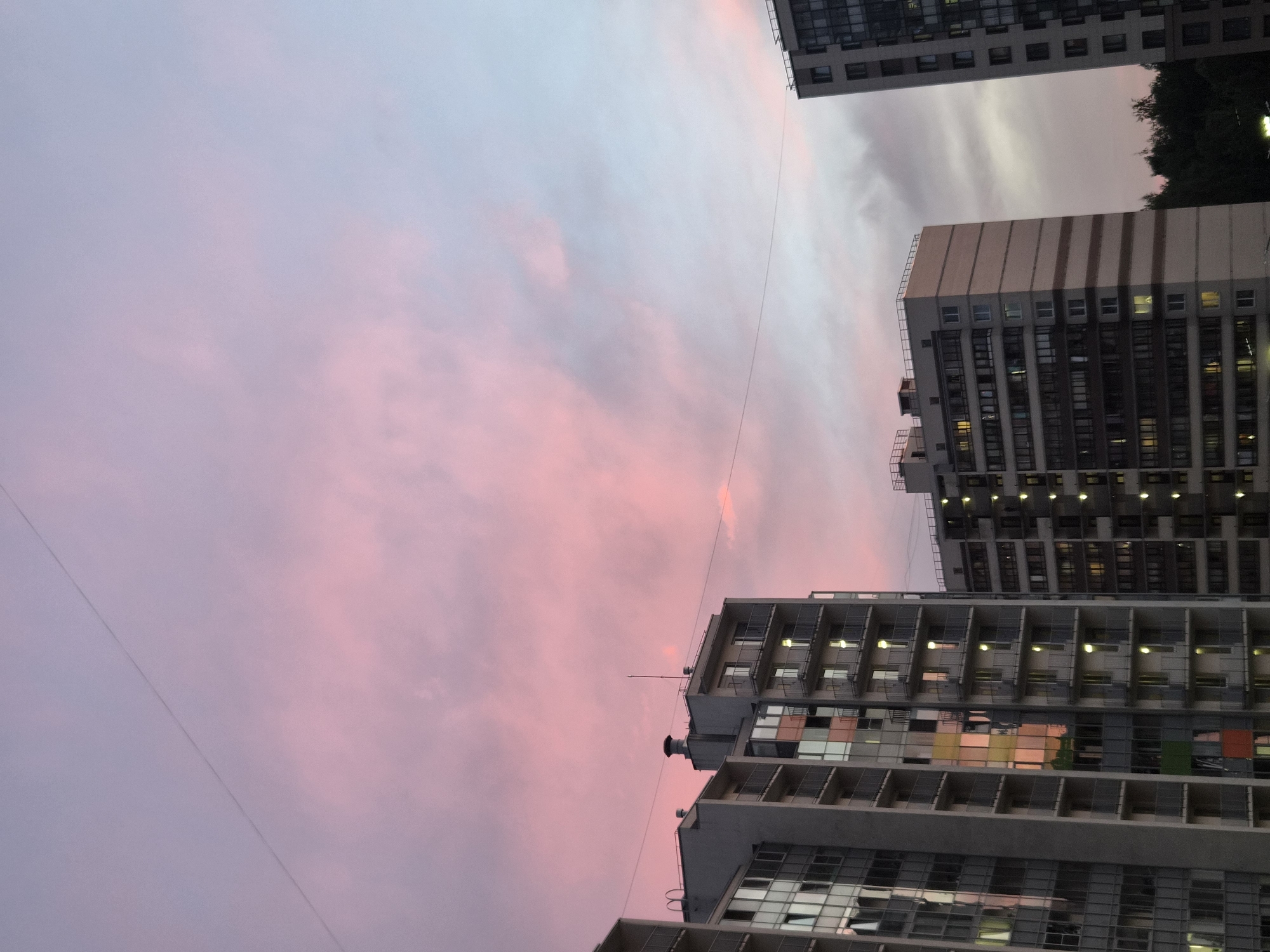 The image depicts a dusk scene in Kudrovo, Russia, characterized by a pastel sky with hues of pink and light blue. Three apartment buildings are visible, with the one on the left being the tallest and featuring a glass facade that reflects the sky's colors. The middle building is a lighter shade with horizontal bands. The building on the far right is also an apartment complex. A thin wire stretches diagonally across the sky, and a line of trees is visible in the background. The lighting suggests it is either early morning or late evening, with lights on in some of the apartment windows. No people are visible in the frame.
