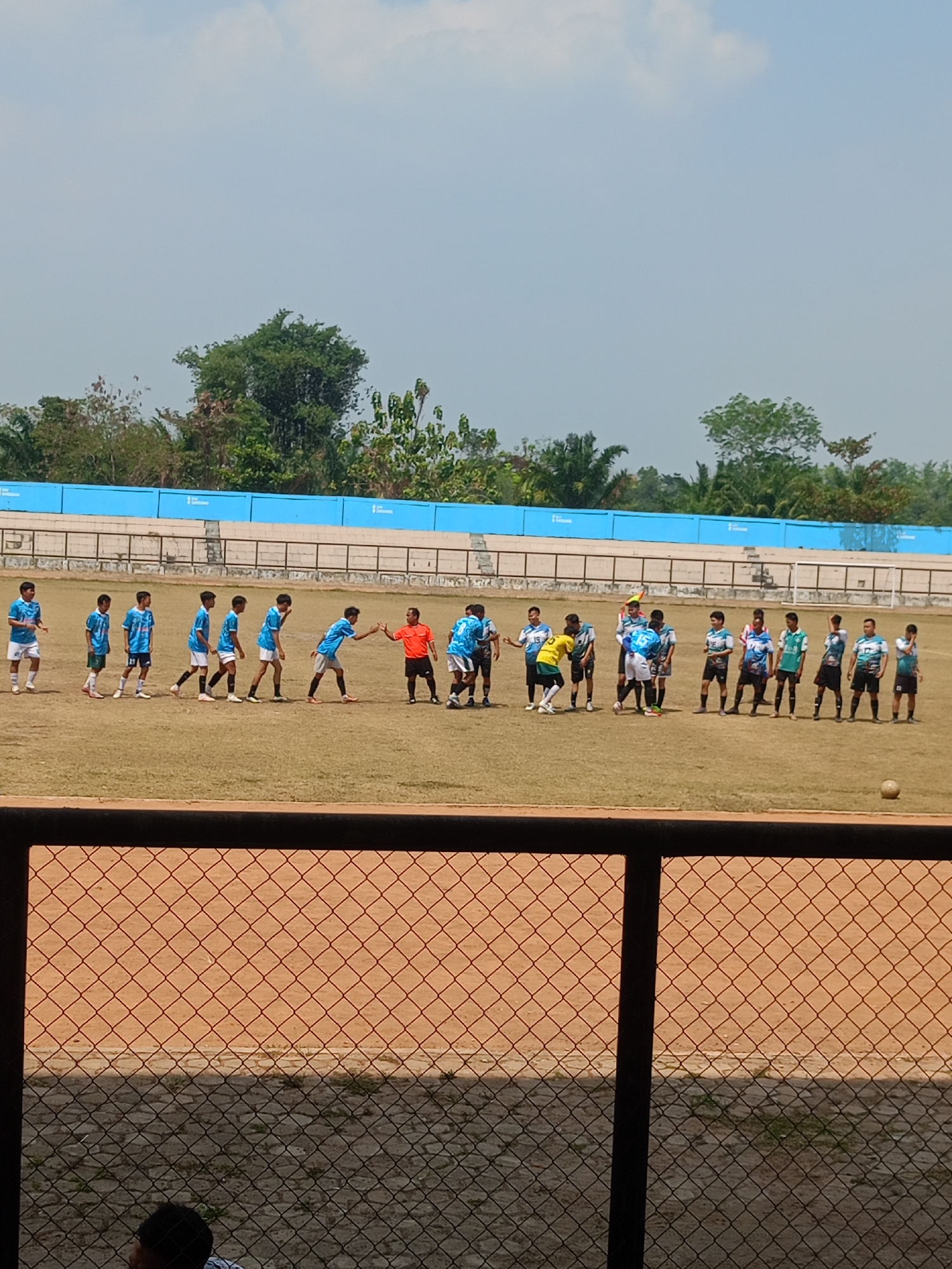 In Prabumulih, Indonesia, a football match is underway on a grassy field with a light brown dirt track surrounding it. The sky is clear and blue with some white clouds. A group of young male athletes, dressed in various colored jerseys and shorts, are positioned on the field, some appearing to be in the middle of a game. One individual in an orange shirt and black shorts, likely a referee, stands among the players. In the background, a blue barrier runs along the edge of the field, with bleachers behind it. Trees and vegetation are visible beyond the stadium structure. A football rests on the field near the foreground. The image is taken from a vantage point behind a fence, partially obscuring the view.