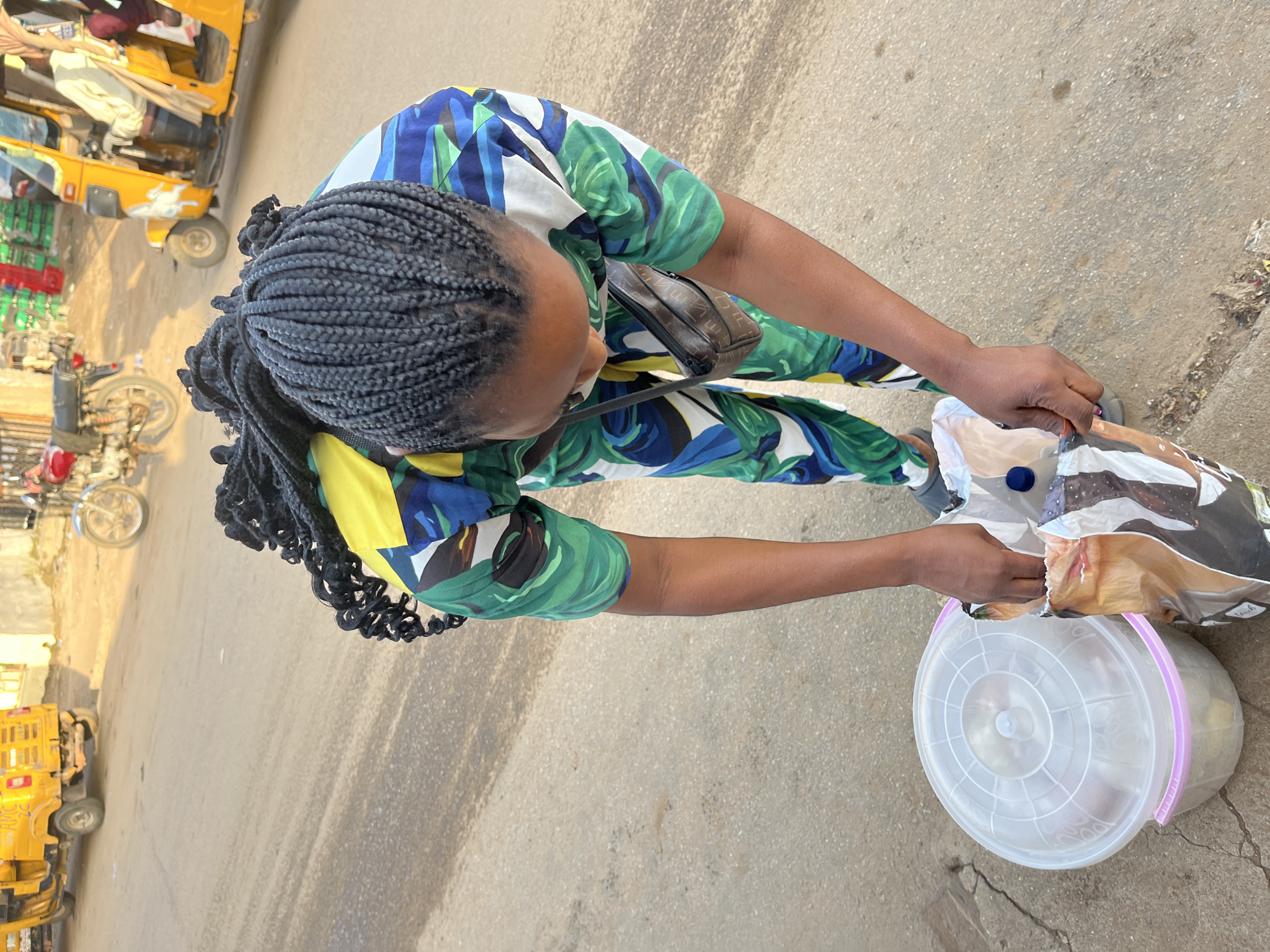 In the city of Abuja, Nigeria, a woman with braided black hair, wearing a green, blue, yellow, and white patterned outfit, is bending down and placing items into a large clear plastic bucket with a pink rim. Beside the bucket, she is also holding open a patterned plastic bag and reaching inside it. A dark brown purse with a strap is slung across her body. The scene takes place on a paved road. In the background, there are two yellow three-wheeled vehicles, one partially visible on the left and a more prominent one on the right with a driver. A motorcycle is also visible further in the background. The environment appears to be an outdoor, urban setting with some debris on the side of the road.