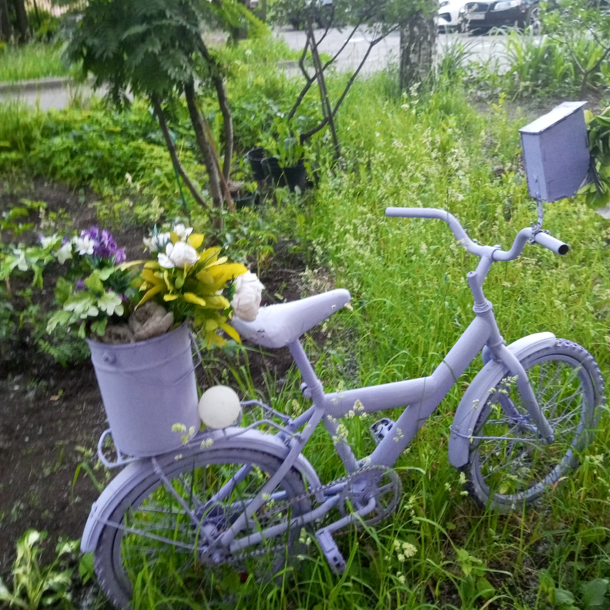 A pastel purple bicycle is parked in a grassy area next to a bucket overflowing with purple and white flowers. The bicycle has a basket attached to the handlebars, and a small box-like structure is mounted behind the seat. The scene appears to be in a park or a garden, with trees and foliage visible in the background. There are no people present in the image. The overall setting suggests a quiet, outdoor space, possibly a garden decoration.