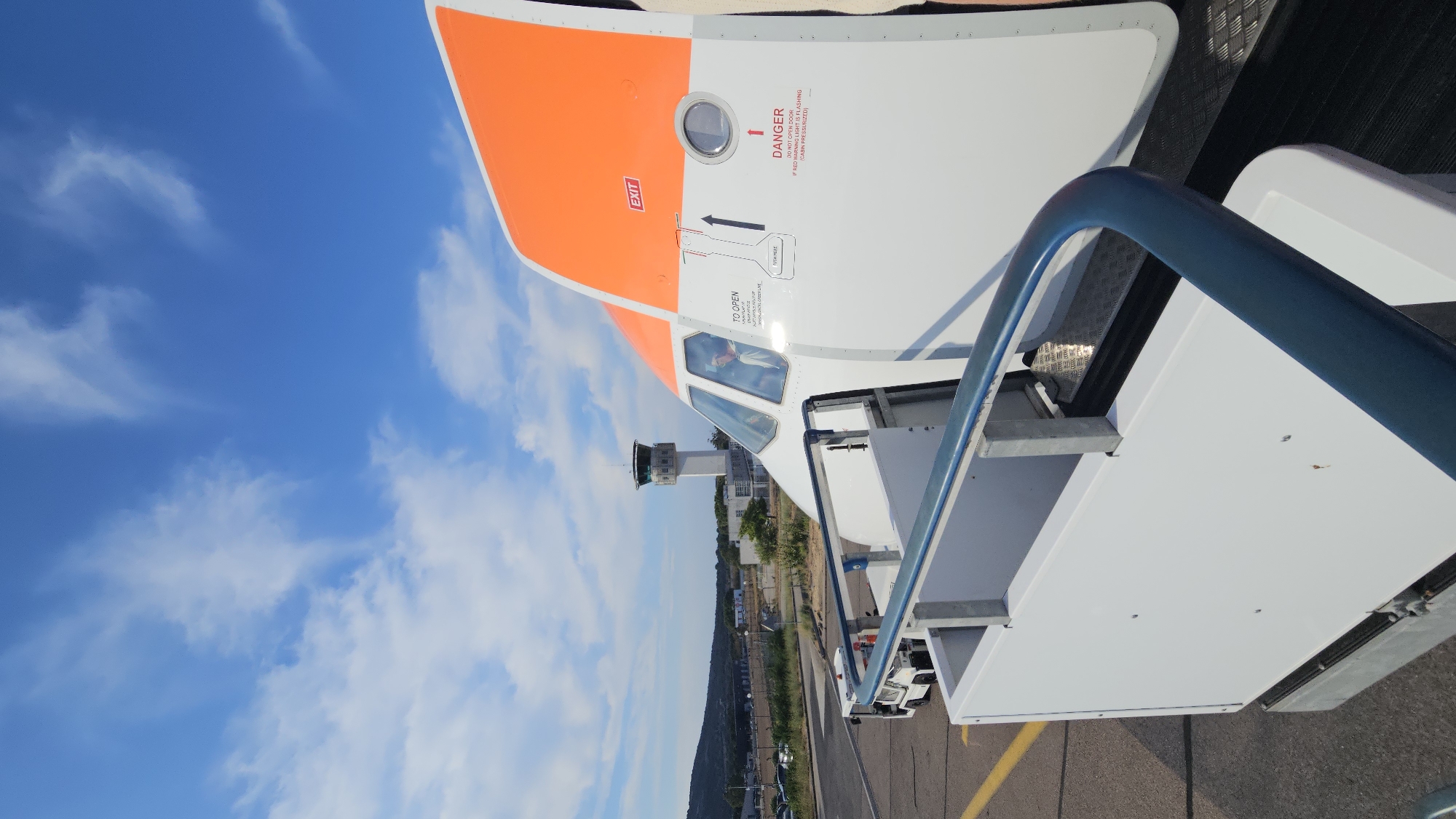 The image shows the side of a passenger airplane, specifically the front section of the fuselage, with an open door and a boarding staircase positioned in front of it. The airplane is white with an orange accent on the top rear of the fuselage. There are various warning labels visible on the side of the aircraft, including an "EXIT" sign and a "DANGER" notice. A pilot or co-pilot can be seen in the cockpit window.
In the background, an airport control tower stands tall, with a building complex and some vegetation behind it. The scene is set on an asphalt tarmac, with yellow lane markings visible. The sky is a clear blue with scattered white clouds. The presence of the airplane, control tower, and tarmac strongly suggests this is an airport environment. Given the context provided in the prompt, this location is the airport of Calvi, France.
There is no indication of any specific actions or interactions taking place other than the airplane being stationary and prepared for boarding or disembarking. The overall impression is one of a typical operational scene at an airport on a clear day.