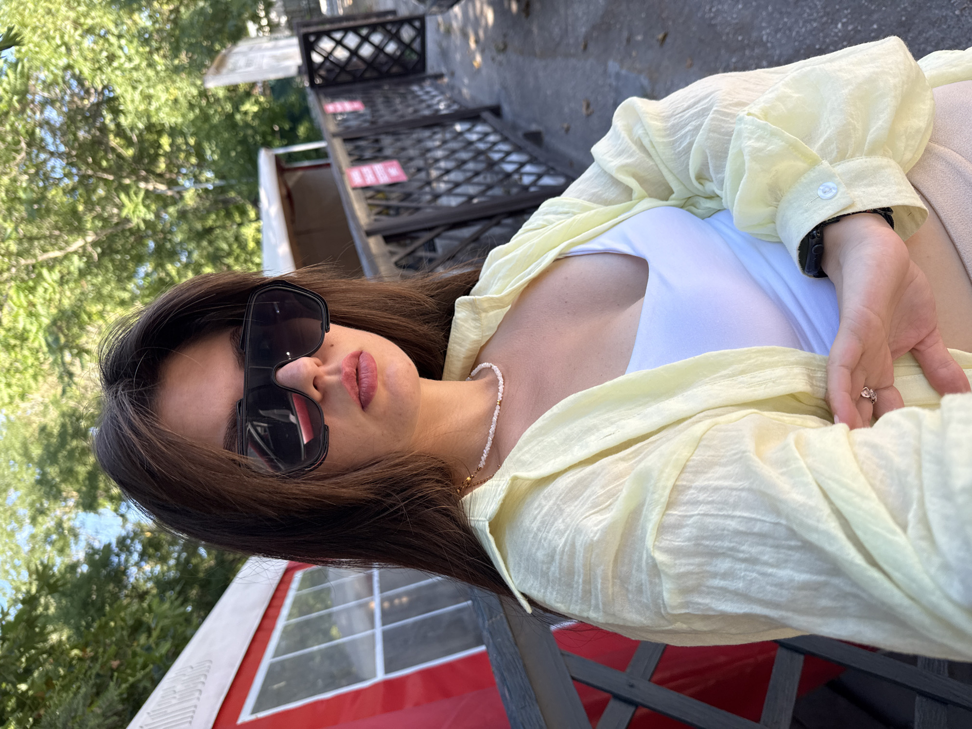 A woman wearing sunglasses and a light yellow open shirt over a white tank top poses for a photo outdoors. She has long dark hair and is wearing a pearl necklace and a ring on her left hand. In the background, there is a red and white structure, a wooden fence, and trees. The setting appears to be a park or an outdoor seating area.