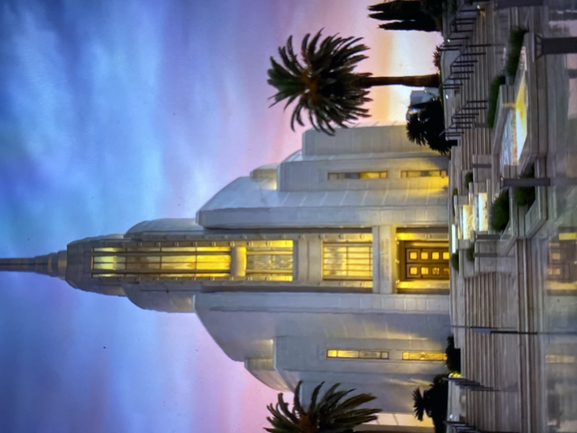 The image depicts the Ciudad de Tinaquillo Venezuela Temple. The temple is a white, multi-tiered Art Deco-style building, illuminated with golden light emanating from its windows and entrance. Palm trees are visible in the foreground and to the side of the building. A plaza with steps and decorative elements leads up to the temple. The sky behind the temple displays a gradient of colors, transitioning from blue to pink, suggesting either sunrise or sunset.