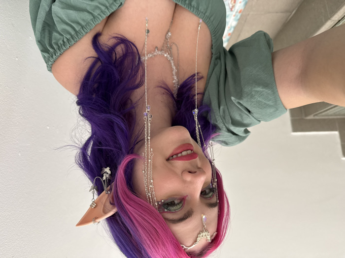 A woman with pointed ears and pink and purple hair faces the camera, smiling faintly. She wears an elaborate silver headpiece and multiple silver chains with embellishments. She sports a green top with gathered sleeves. The background includes a plain white wall and a glimpse of a tiled floor. The image is oriented sideways.