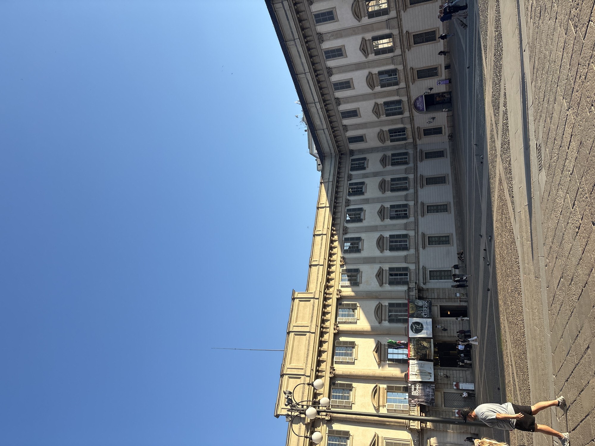 This image depicts a sunny day in a European city, identifiable as Milan, Italy, with a large, classical building dominating the right side of the frame and a clear blue sky filling the left. The multi-story building features a light-colored facade with numerous symmetrically placed windows, many equipped with dark shutters. Architectural details like cornices and decorative elements are visible.
Hanging from the building's facade are several banners and posters advertising cultural events and institutions. Visible text includes "KIEFER" (likely referring to artist Anselm Kiefer), "MUSEO DIOCESANO," "DANTE," "DUOMO," "ART EXHIBITION," and "GALLERIA VITTORIO," providing strong location cues related to Milan's cultural landmarks. A slender antenna or spire extends from the building's roof upwards into the sky.
The foreground and left side of the scene show a wide, paved public square or street. The paving consists of cobblestones and patterned gray sections. Bright sunshine casts distinct shadows, suggesting the time of day is late morning or early afternoon. Several people are present, engaged in pedestrian activity. In the immediate foreground, a man wearing a grey t-shirt, black shorts, and sneakers, along with a baseball cap, walks away from the camera. Other individuals are seen further back, near the building's entrances. A black lamppost with multiple globe lights stands in the lower part of the scene, next to the building. A few pigeons are scattered on the ground. The overall atmosphere is that of a lively urban setting on a clear, warm day.
