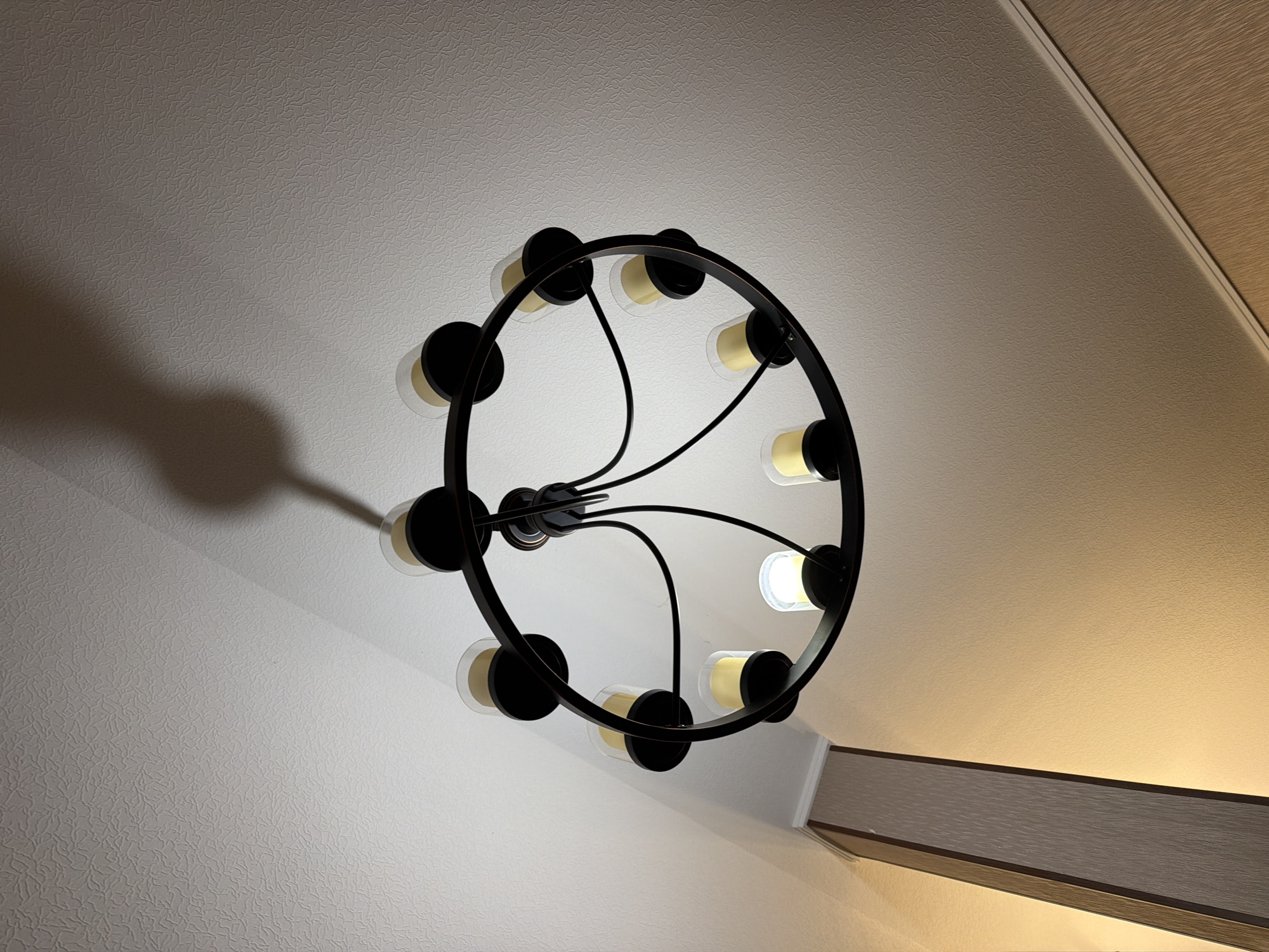 A dark metal chandelier with multiple candle-shaped lights hangs from a textured white ceiling in Koshkurgon, Uzbekistan. The chandelier has a circular frame from which eight arms extend, each holding a light. Some lights are illuminated, appearing as yellow flames within clear glass shades, while others are unlit. A distinct shadow of the chandelier is cast on the ceiling. To the left of the chandelier, a portion of a wall is visible, featuring a vertical wooden trim with a textured surface. The overall lighting in the scene is dim, with the chandelier being a primary light source.