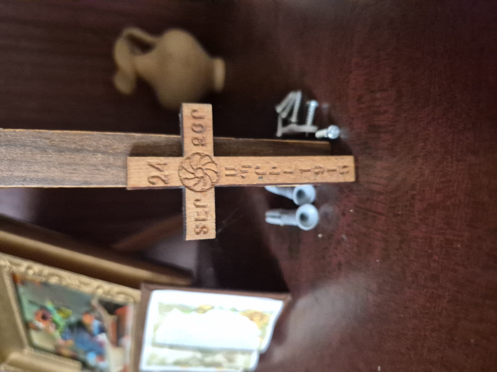 A close-up, medium-angle, eye-level shot shows a wooden cross with inscribed text and a decorative element. The cross is positioned vertically and has a horizontal beam. The text on the cross appears to be "SEP" on the left, a circular symbol with radiating lines in the center, and "BOR" on the right. Below the horizontal beam, vertically inscribed text is partially visible, reading "URC". Further down, another line of text is visible, with numerals "1915". To the left of the cross, in the blurred background, a framed picture with colorful art is visible. To the right of the cross, a blurred, light-colored vase-like object is present. In the foreground, directly below the cross, are several small, grey, wall anchors and some metallic screws, suggesting the cross might be intended for wall mounting. The surface beneath the objects is a dark, reddish-brown, polished wood, likely a table or shelf. The lighting is soft, casting subtle shadows. The overall scene is still and domestic, with no people present. The location is not specified in the image.