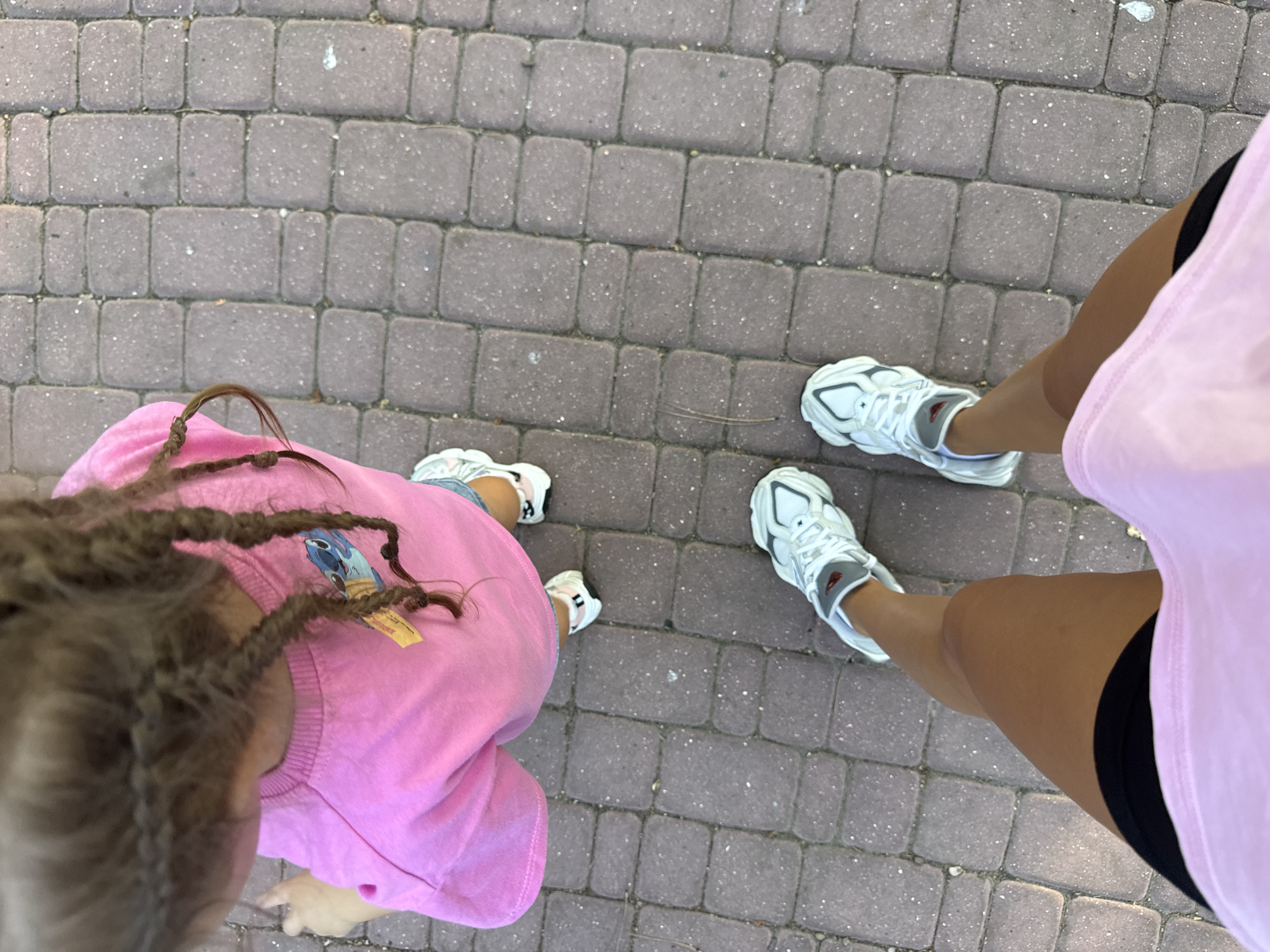 The image shows the legs of two people standing on a brick-paved pathway. Both individuals are wearing white athletic shoes. The person on the left is wearing a bright pink sweatshirt, and their braided hair is visible. The person on the right is wearing a light pink t-shirt and black shorts, with one leg in the foreground. The scene appears to be outdoors, possibly in a park or a pedestrian area, given the brickwork and the lighting suggesting daytime. The overall impression is one of casual activity, perhaps walking or taking a break. There are no discernible emotional cues or specific weather indicators, but the lighting suggests it's a bright day. No visible text is present in the image. The location context of Chișinău, Moldova, does not provide specific visual cues within the image itself.