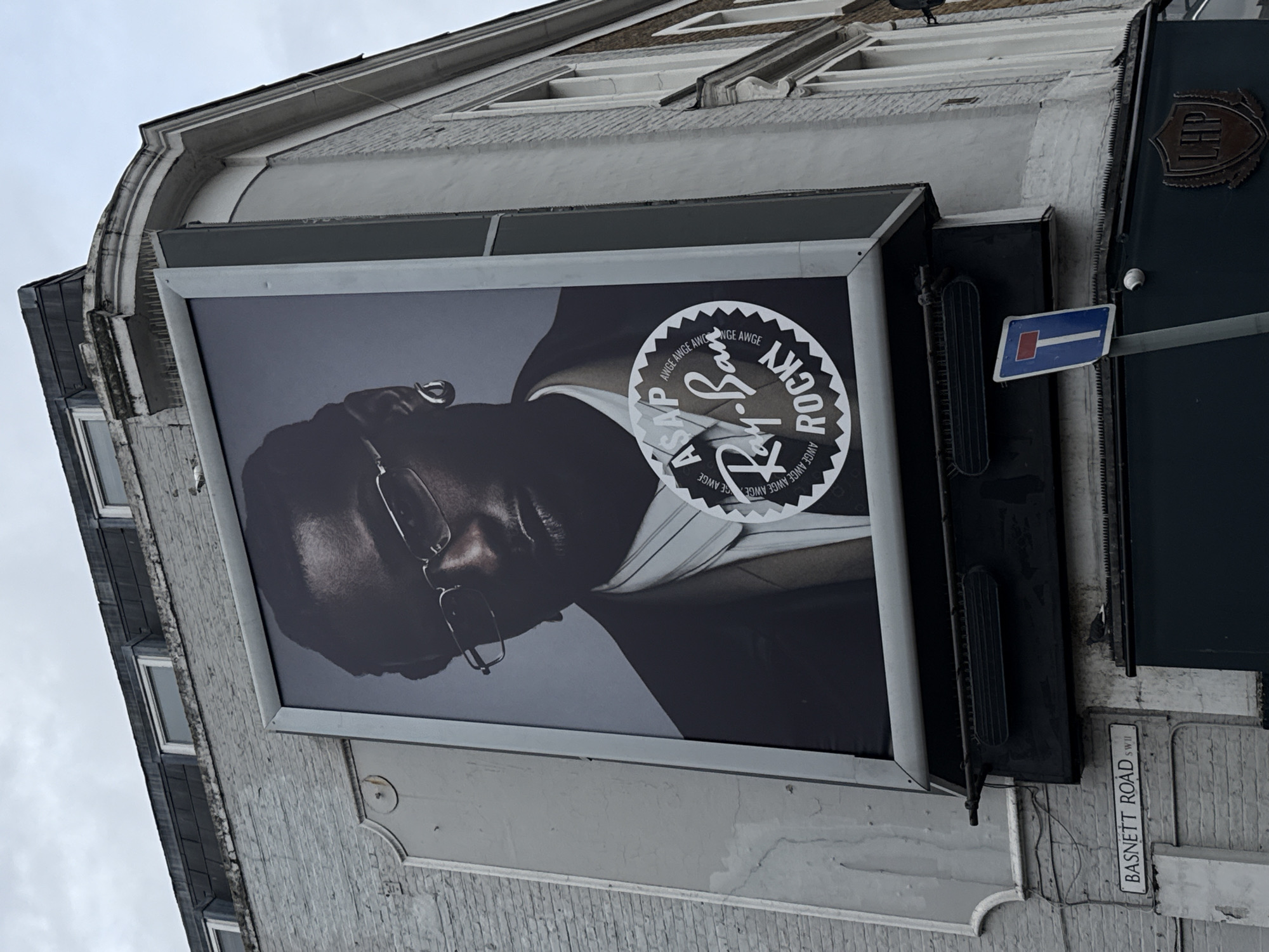 The image depicts an advertisement for Ray-Ban featuring ASAP Rocky on a building in London, United Kingdom. The advertisement is prominently displayed on a gray building with visible architectural details, including windows and decorative elements. The Ray-Ban logo and ASAP Rocky's name are visible on the ad. A "No Through Road" sign and a sign indicating "Basnett Road SW11" are adjacent to the advertisement. The scene is captured from a low angle, emphasizing the height of the building and the size of the advertisement.