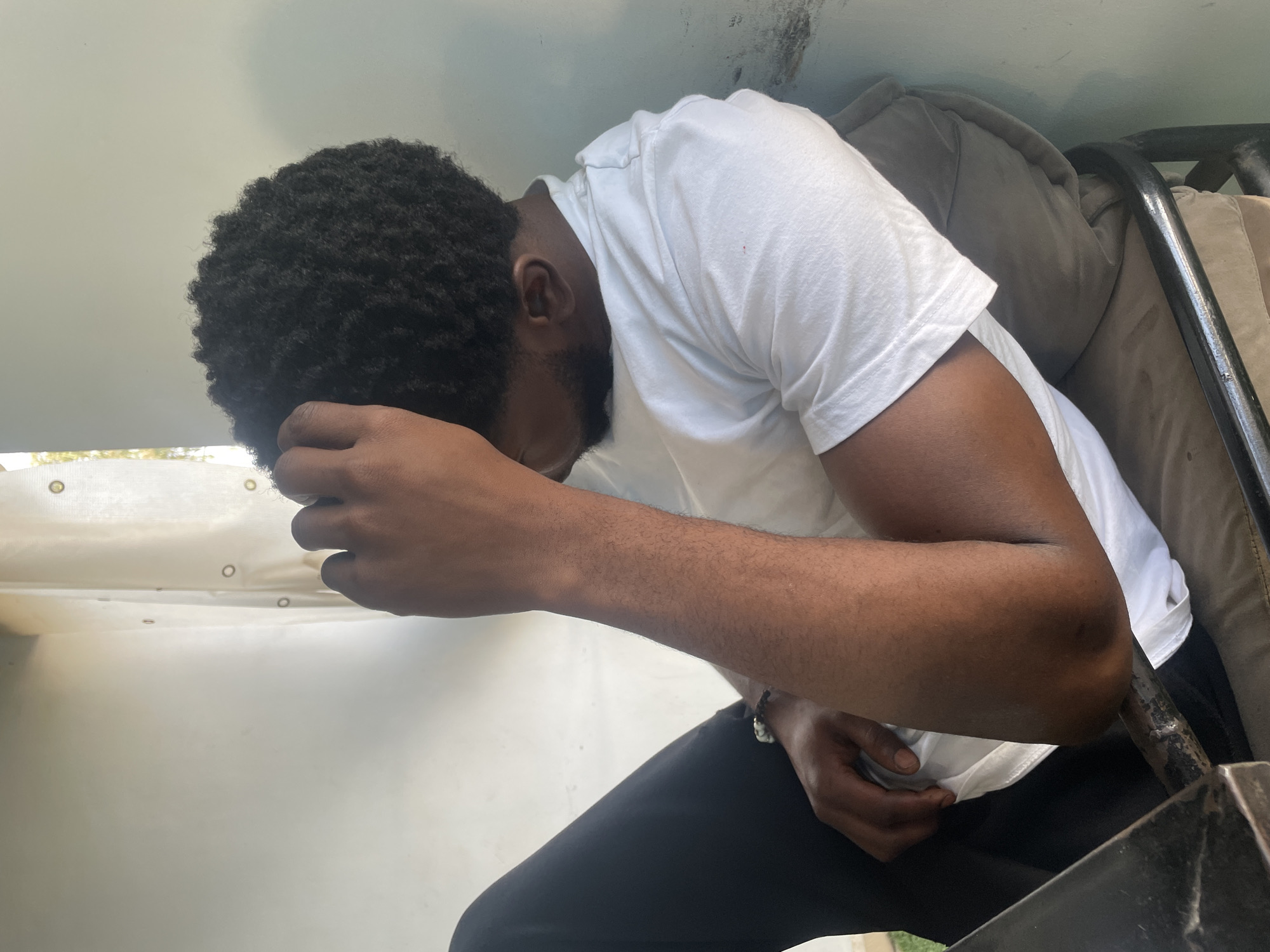 A dark-skinned man with a beard and afro is seated with his head in his hand. He is wearing a white t-shirt and dark-colored pants. He appears to be indoors, next to a white wall. A seat is visible to his right. The location of the image is unknown.