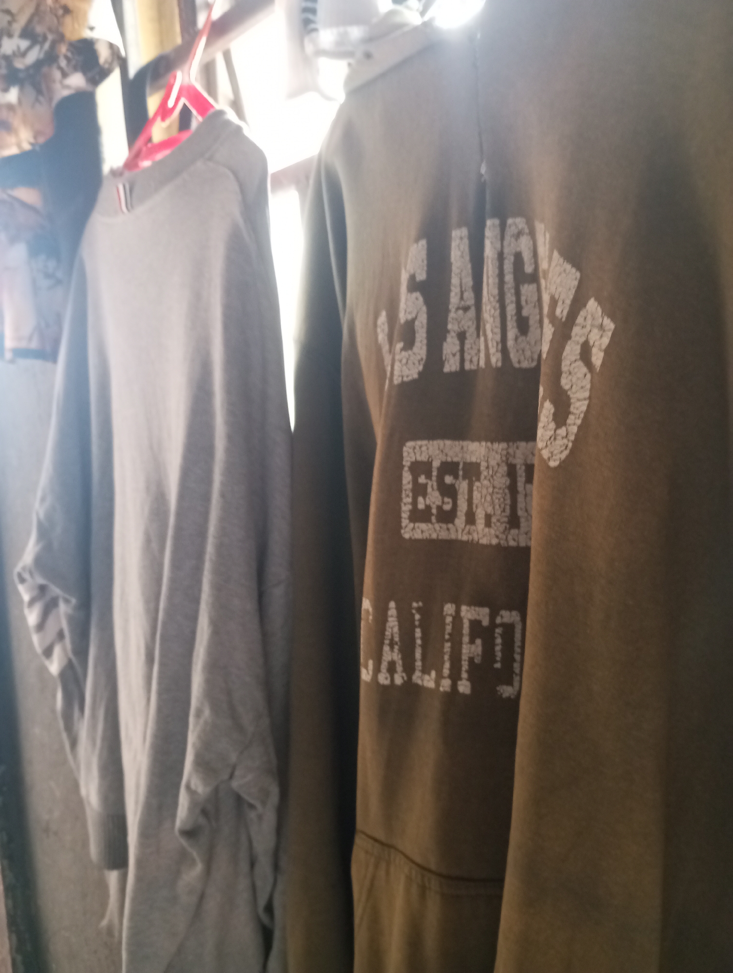 The image shows two garments hanging on a clothesline. On the left is a light grey long-sleeved shirt, possibly a sweatshirt, with a portion of a white striped cuff visible. On the right is a dark olive green hoodie with white lettering that reads "LOS ANGELES EST. 1949 CALIF." The garments are hanging in what appears to be an indoor or semi-outdoor space with a window or opening in the background letting in bright light. The context of Onitsha, Nigeria is not discernible from the visual elements present in the image.