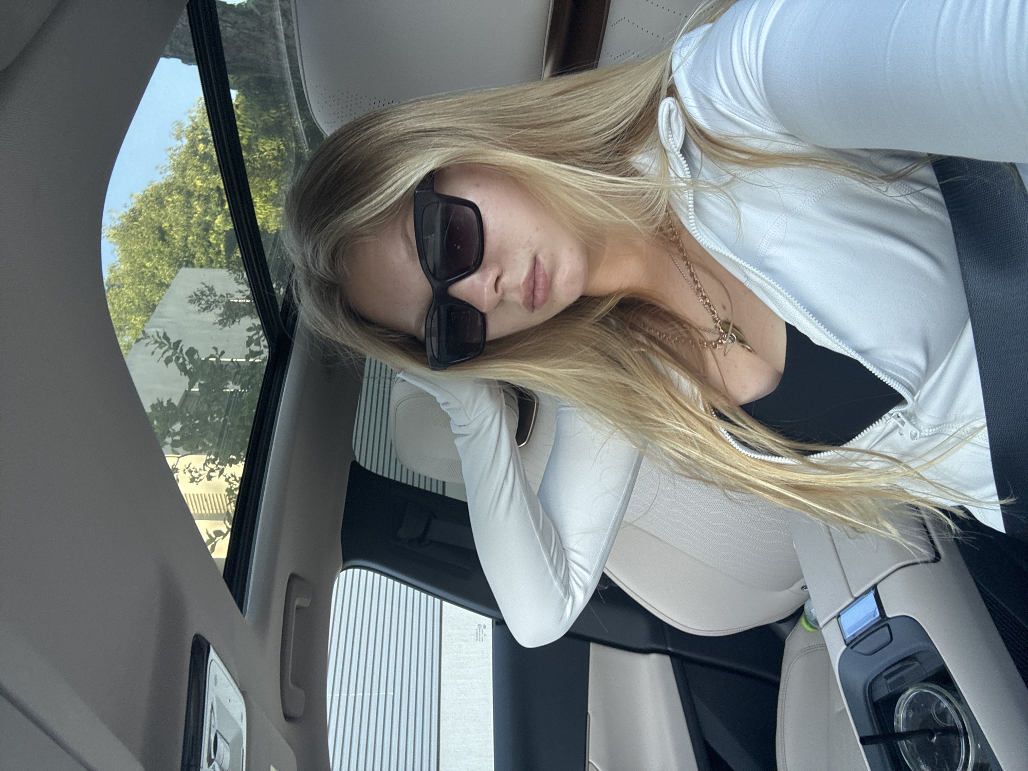 A young woman with long, straight blonde hair is captured in a selfie or portrait, seated in what appears to be the passenger seat of a car. She is looking directly at the camera with a neutral expression, her eyes concealed by large, rectangular black sunglasses. She wears a white long-sleeved athletic top with a partial zipper, revealing a black top underneath, and a delicate gold necklace with a pendant. A dark seatbelt crosses her chest. Her left arm, covered by the white sleeve, is bent and rests comfortably against the car's light-colored interior.
The car's interior features light-toned upholstery, possibly leather, on the seats and ceiling. Through the car windows, lush green trees and a clear blue sky are visible, indicating bright daylight and good weather. Elements of the car's sunroof and a dark frame around the window are also seen. On the center console, a clear beverage cup with a straw is present. The overall setting is inside a modern vehicle, suggesting a journey or a stopped car. No discernible text is visible within the image. The location context provided is Kfar Shmaryahu, Israel.