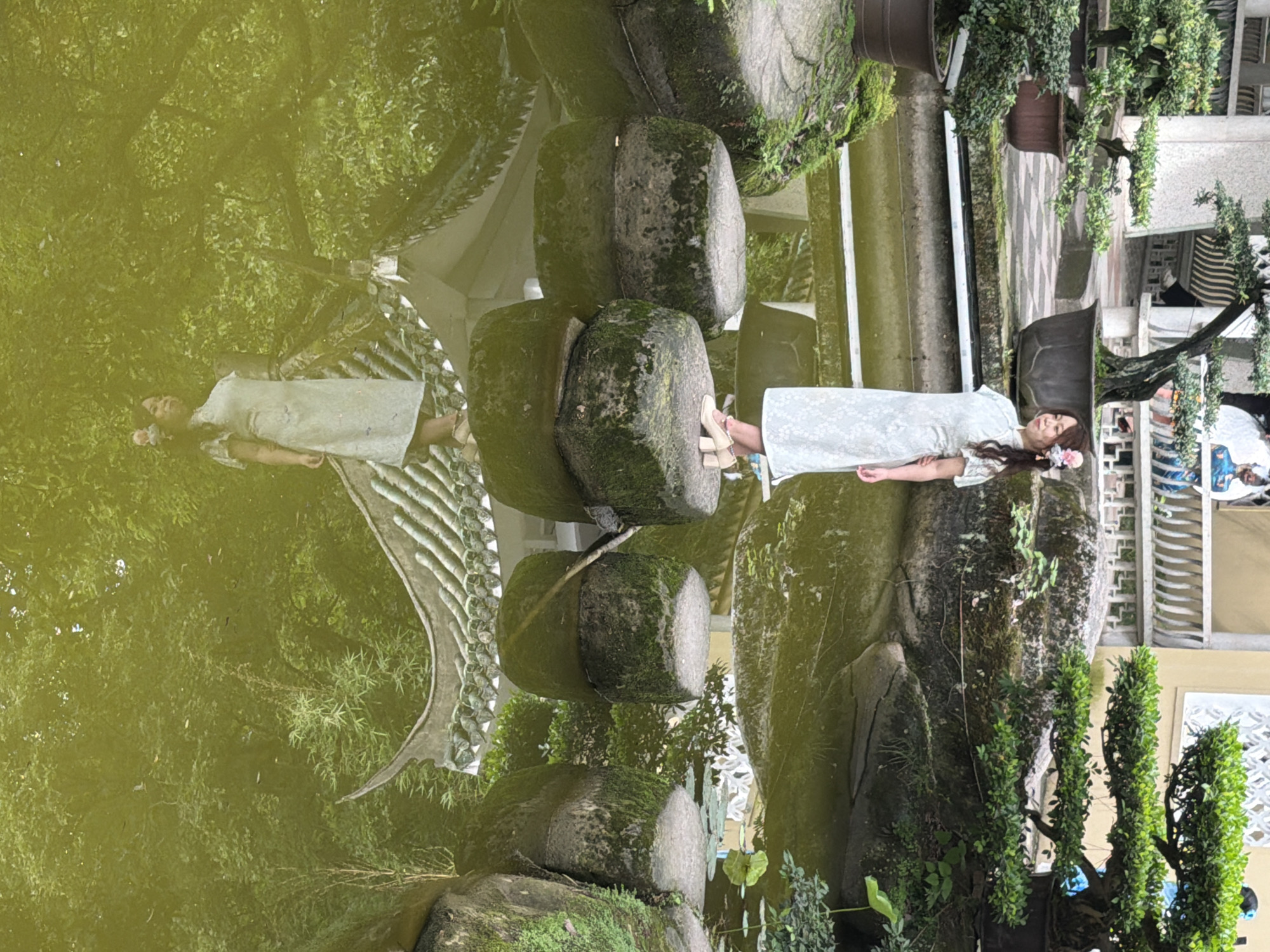 A woman stands on stepping stones in a pond surrounded by bonsai trees. She is wearing a light blue dress and has flowers in her hair. The pond water is green and reflects the surrounding trees and a decorative bridge. The scene appears to be a garden or park.