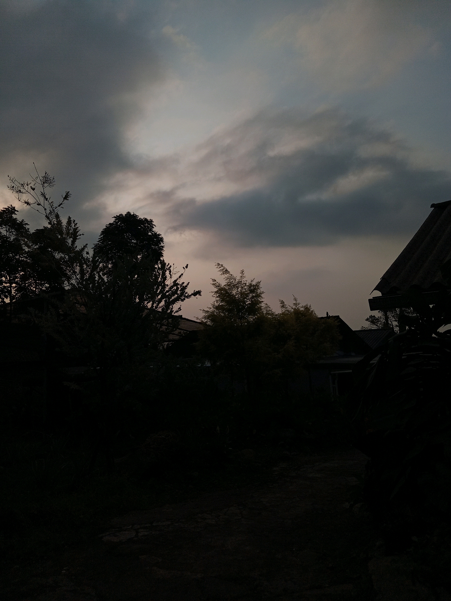 The image depicts a dark, overcast sky over a silhouetted landscape. In the foreground, dense vegetation and what appear to be the roofs of houses are visible, almost entirely consumed by shadow. The sky is a gradient of grays and muted blues, with some lighter patches suggesting the presence of clouds. A portion of a corrugated roof, likely from a residential structure, juts into the frame from the right. The overall impression is of a rural or semi-rural setting at dusk or under heavy cloud cover. The location is identified as Claket, Indonesia.