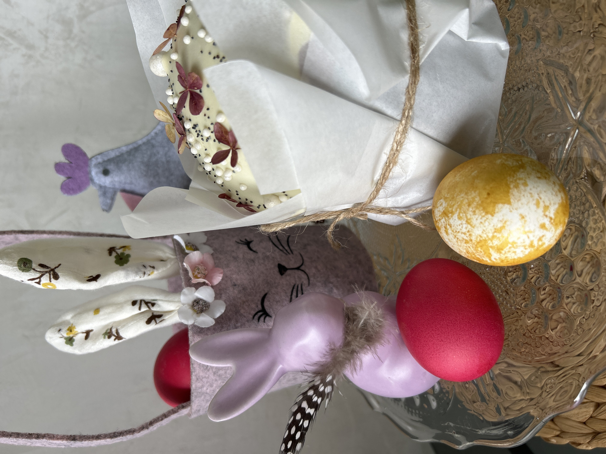 The image shows an Easter-themed arrangement on what appears to be a glass plate with a woven base. The central element is a white chocolate bark decorated with dried flowers and white pearl-like candies, wrapped in white parchment paper and tied with twine. Next to it are dyed Easter eggs in red and yellow. There's also a felt bunny head with floral ears and a small ceramic bunny figurine, with a feather placed near the ceramic bunny. A felt bird-shaped decoration is visible beside the wrapped chocolate. The setting appears to be indoors. There is no information in the picture to suggest location.