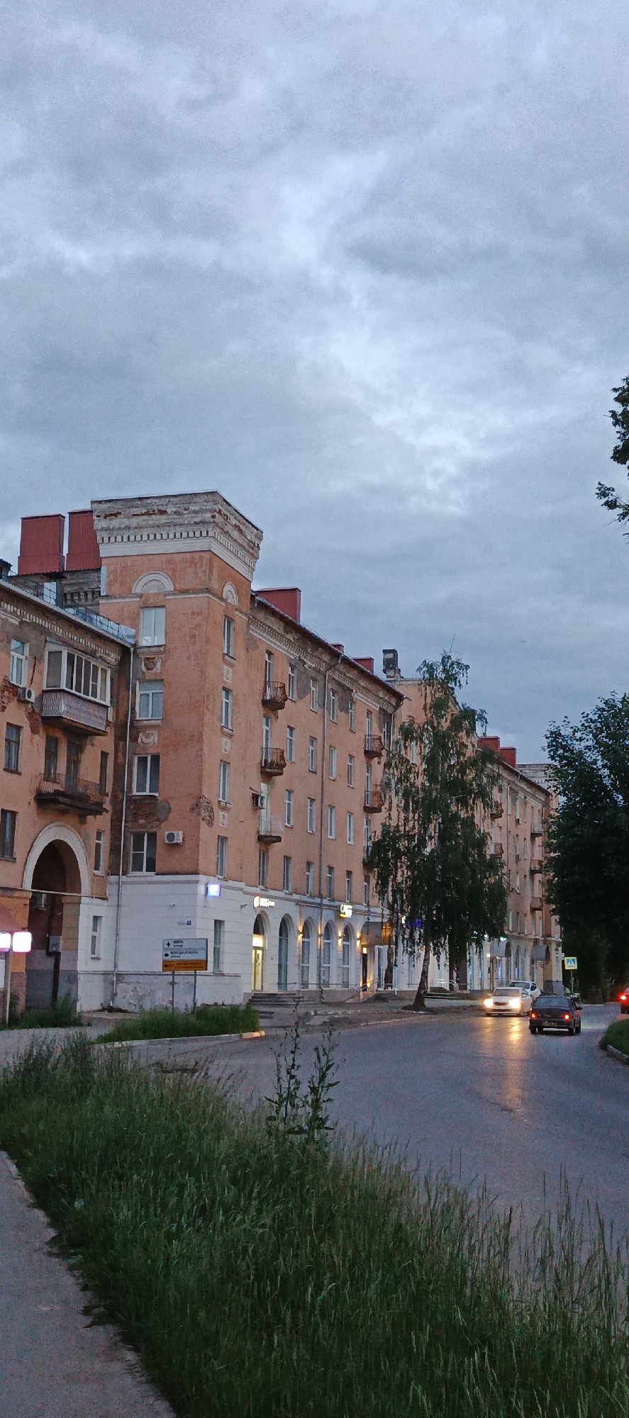 The image shows a street scene in Votkinsk, Russia, at dusk. An old, multi-story apartment building with peach-colored facades and red-tiled roofs lines the street on the left. The building features arched ground-floor entrances and balconies. Several cars are driving on the wet asphalt road, their headlights illuminating the scene. Tall, green trees stand on the right side of the road, partially obscuring the view of another building. The sky is overcast with gray clouds. Green grass and weeds grow along the side of the road and sidewalk in the foreground. A street sign with Russian text and directional arrows is visible near the apartment building.