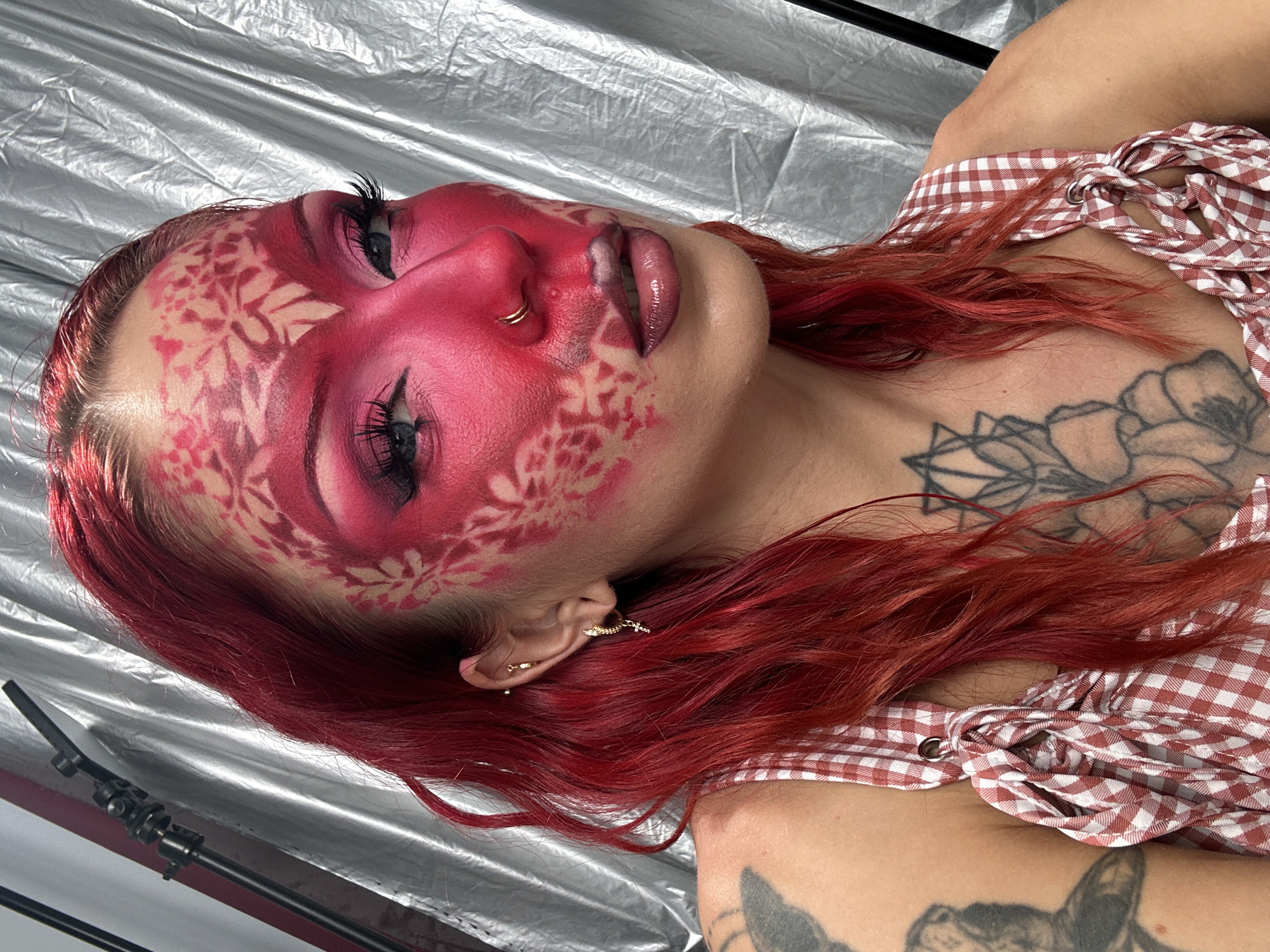 The image shows a close-up, high-angle shot of a person with bright red hair and dramatic pink and red face makeup, including stenciled floral patterns on their forehead and cheeks. The person is wearing a red and white gingham top and has tattoos visible on their shoulder and chest. They are looking towards the camera with a slight tilt of their head. The background is a silver, crinkled fabric backdrop. There are no discernible elements in the image that would suggest a location in North Kuta, Indonesia, or any urban environment.