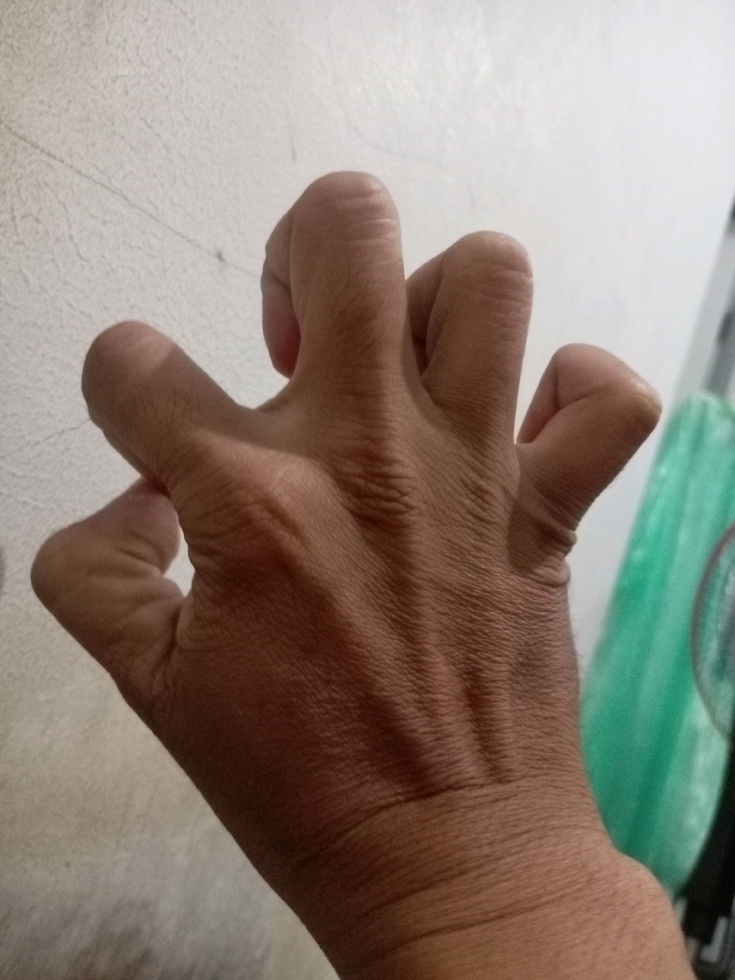 A close-up, medium shot shows the dorsal side of a human hand with fingers contorted into a claw-like position. The skin is tan and appears wrinkled, particularly on the knuckles and between the fingers. The background is out of focus, showing a textured, off-white wall on the left and a blurry green object, possibly a plastic container, on the right. The lighting suggests an indoor environment. The image does not contain any identifiable landmarks that would place it within Batam, Indonesia.