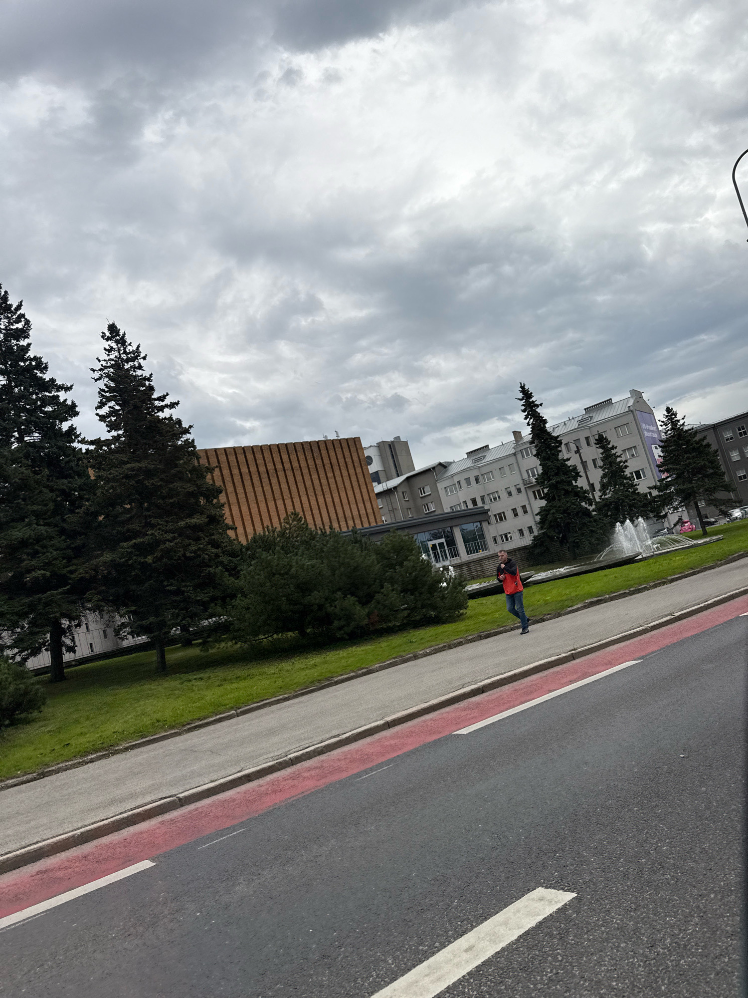 This image depicts a street scene in Tallinn, Estonia, on a cloudy day. In the foreground, a road with a red and white painted lane divider is visible, angled from the bottom right towards the top left. A sidewalk runs alongside the road, separated by a curb. On the sidewalk, a person is walking from left to right. The person is wearing a red vest over a dark shirt and blue jeans.
Behind the sidewalk and a grassy area, several evergreen trees are prominent. To the right of the trees, a fountain with water spraying upwards is situated in a circular basin, surrounded by greenery. Several buildings of varying architectural styles are visible in the background. One building to the left has a distinctive facade with vertical brown wooden or brick elements. Further back, light-colored multi-story buildings with numerous windows are present. The sky is heavily overcast with dark, diffused clouds. A street light pole is partially visible on the far right edge of the frame.