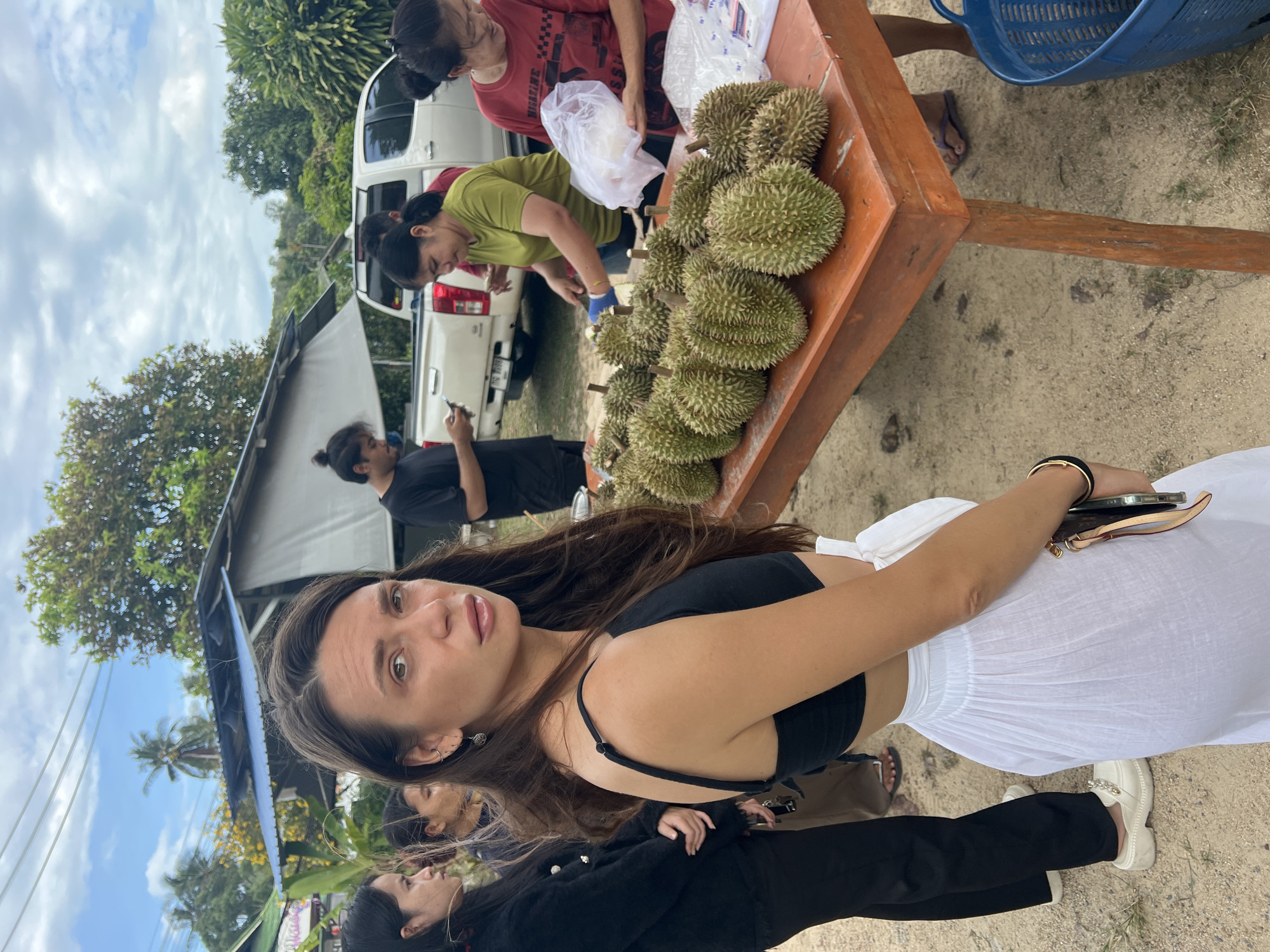 The image shows an outdoor scene in บ้านบางรักษ์, Thailand. In the foreground, a woman with long brown hair, wearing a black top and white skirt, is posing for the camera. Her arm holds a phone. Behind her and to the left, two other women are visible; one wearing a black blazer, and the other is partially obscured.
In the background, there is a fruit stall selling durian. A woman in a green shirt is handling the durians on an orange table, while another woman in a red shirt stands beside her. Further back, a man in a black shirt is looking at his phone next to a white pickup truck. The stall is partially covered by a makeshift roof. Trees are visible in the distance. The ground appears to be sandy.