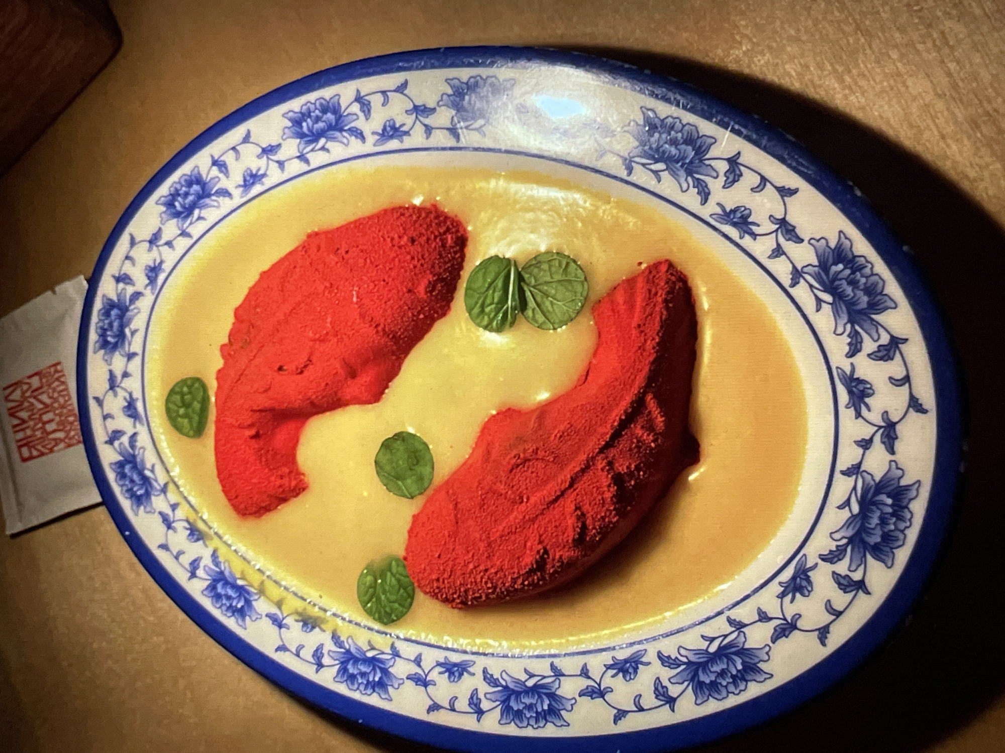An overhead shot captures an oval-shaped plate, white with a blue floral pattern around the rim, containing what appears to be a dish. Two red, roughly fish-shaped mounds lie in a light yellow, sauce-like liquid. Small, round, green mint leaves are sparsely placed around the plate as garnish. The plate is set on a wooden surface, and a portion of a white packet with red lettering, possibly a condiment or seasoning package, is visible on the left side of the frame.